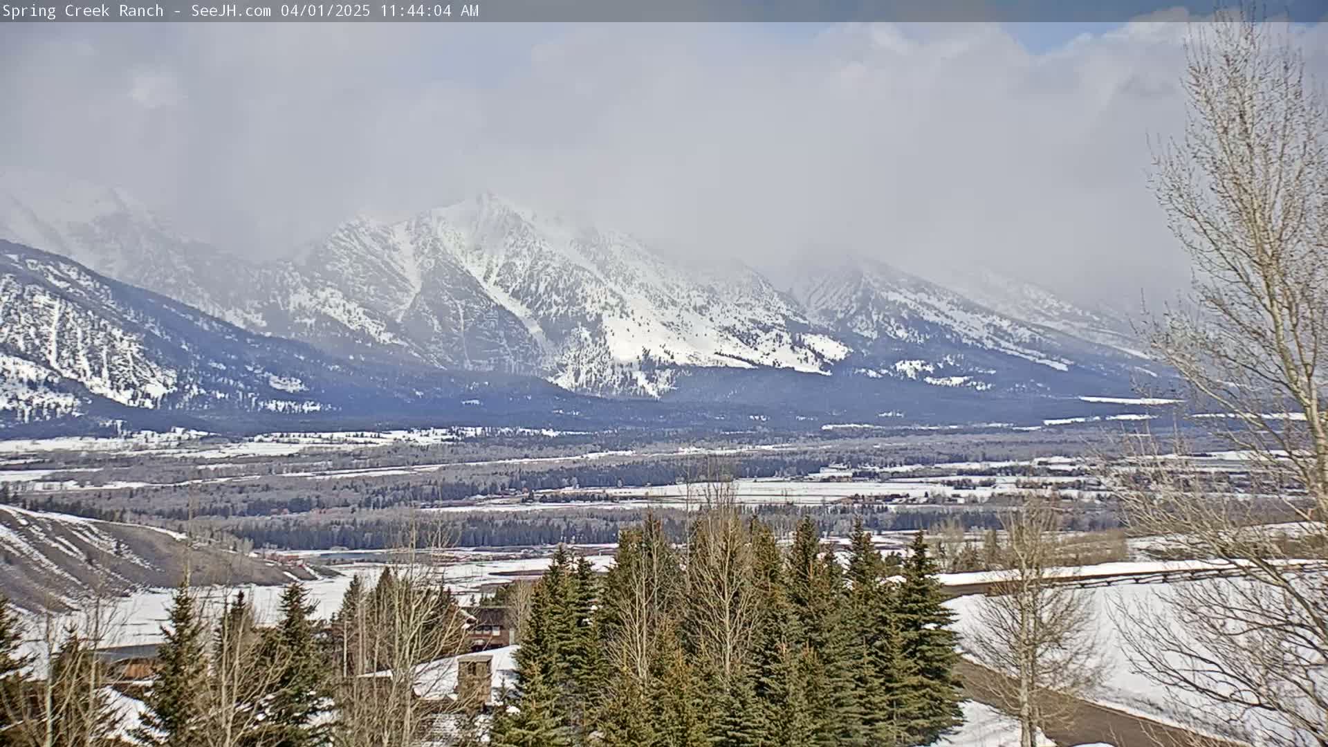 A hazy, sunny day reveals a snow-covered mountain range in the distance, with a valley of snow-dusted trees and buildings in the foreground.