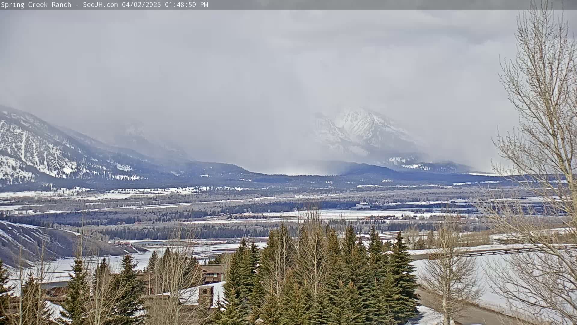 A snow-covered valley with a distant, snow-capped mountain range is visible under a hazy, overcast sky.