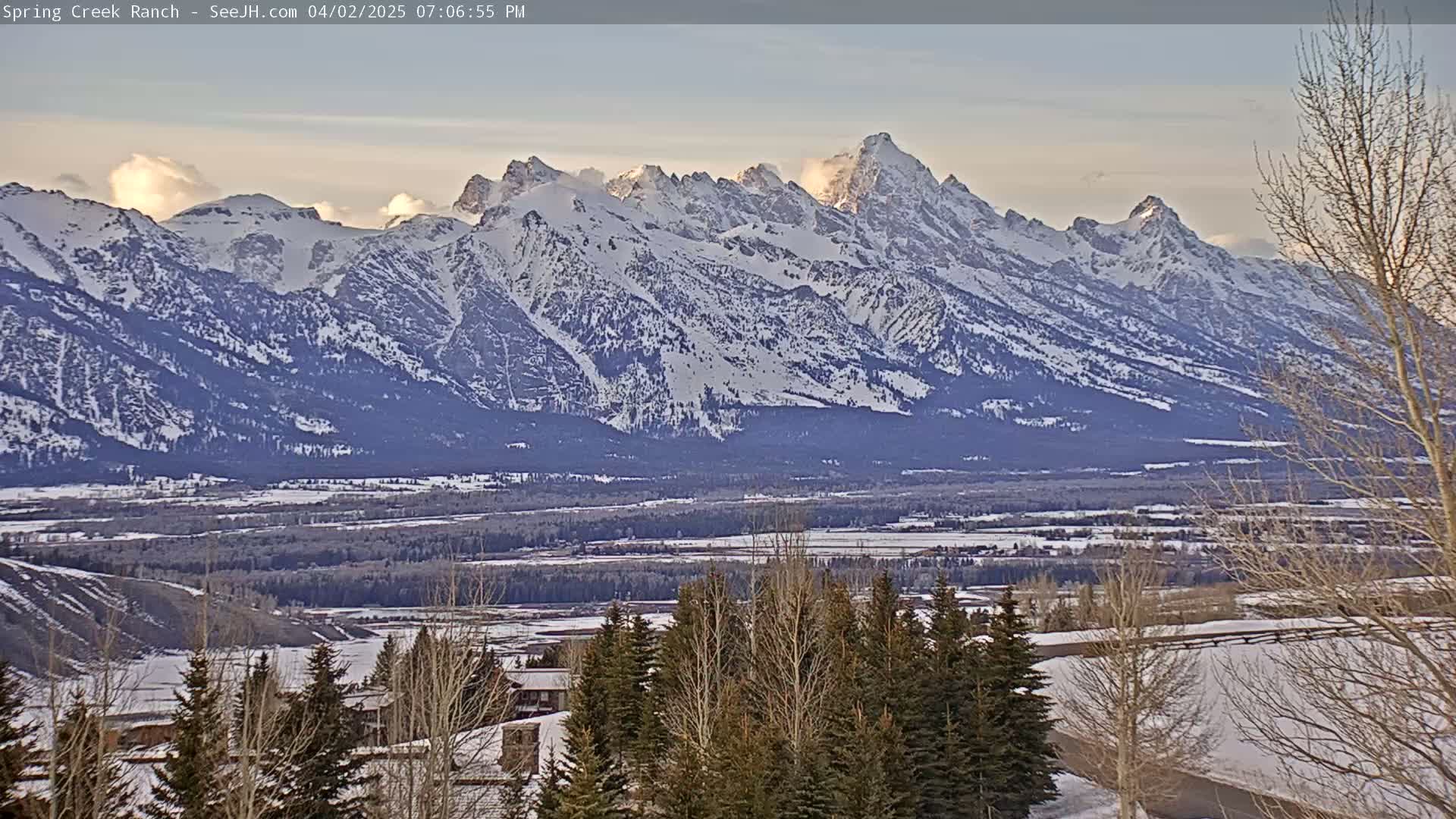 A snow-covered mountain range is visible at sunset, with a valley of snow-covered trees and buildings in the foreground.