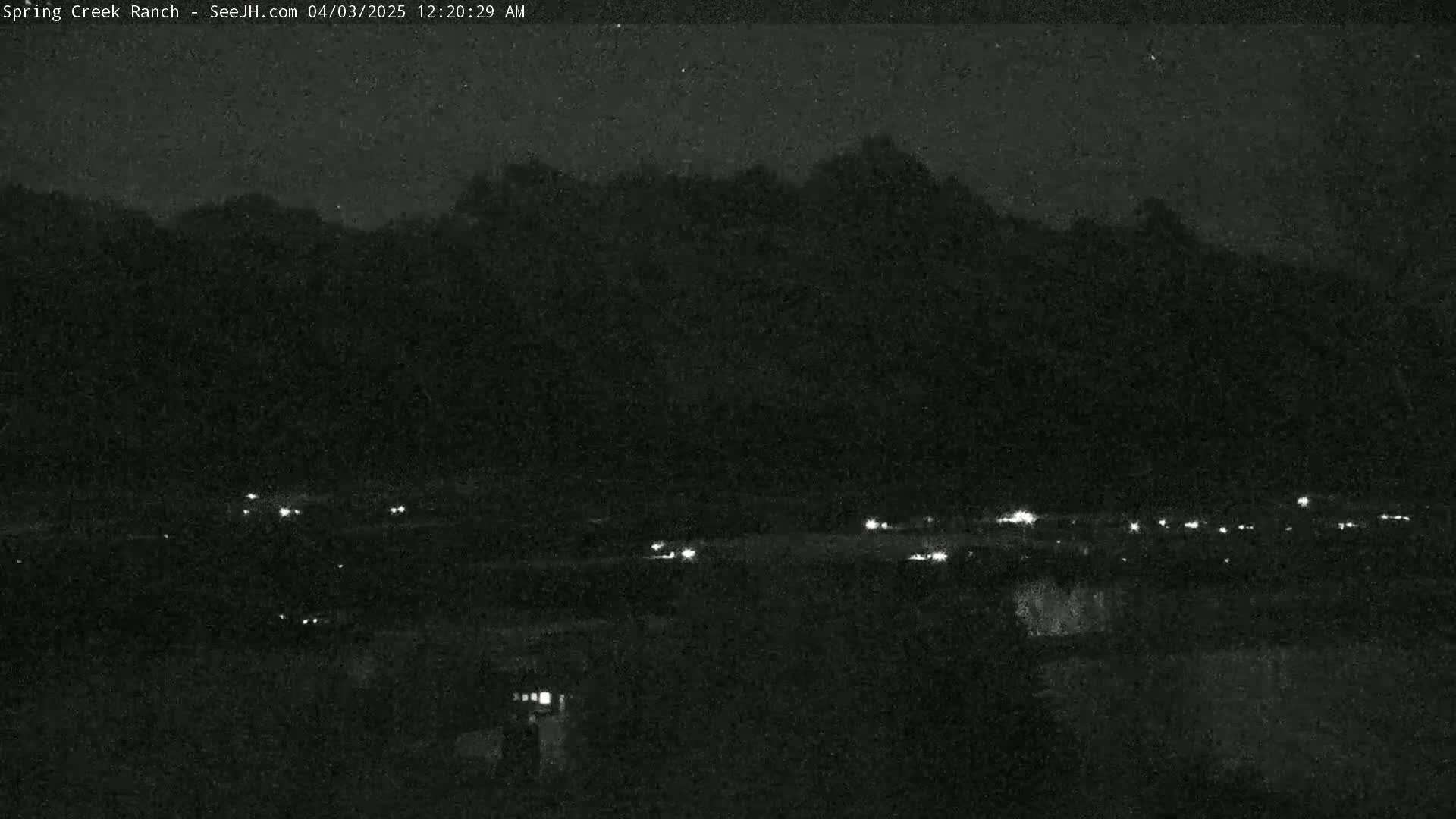A nighttime long shot shows a dark landscape with a few scattered lights in the foreground and a dark mountain range silhouetted against a mostly clear night sky.