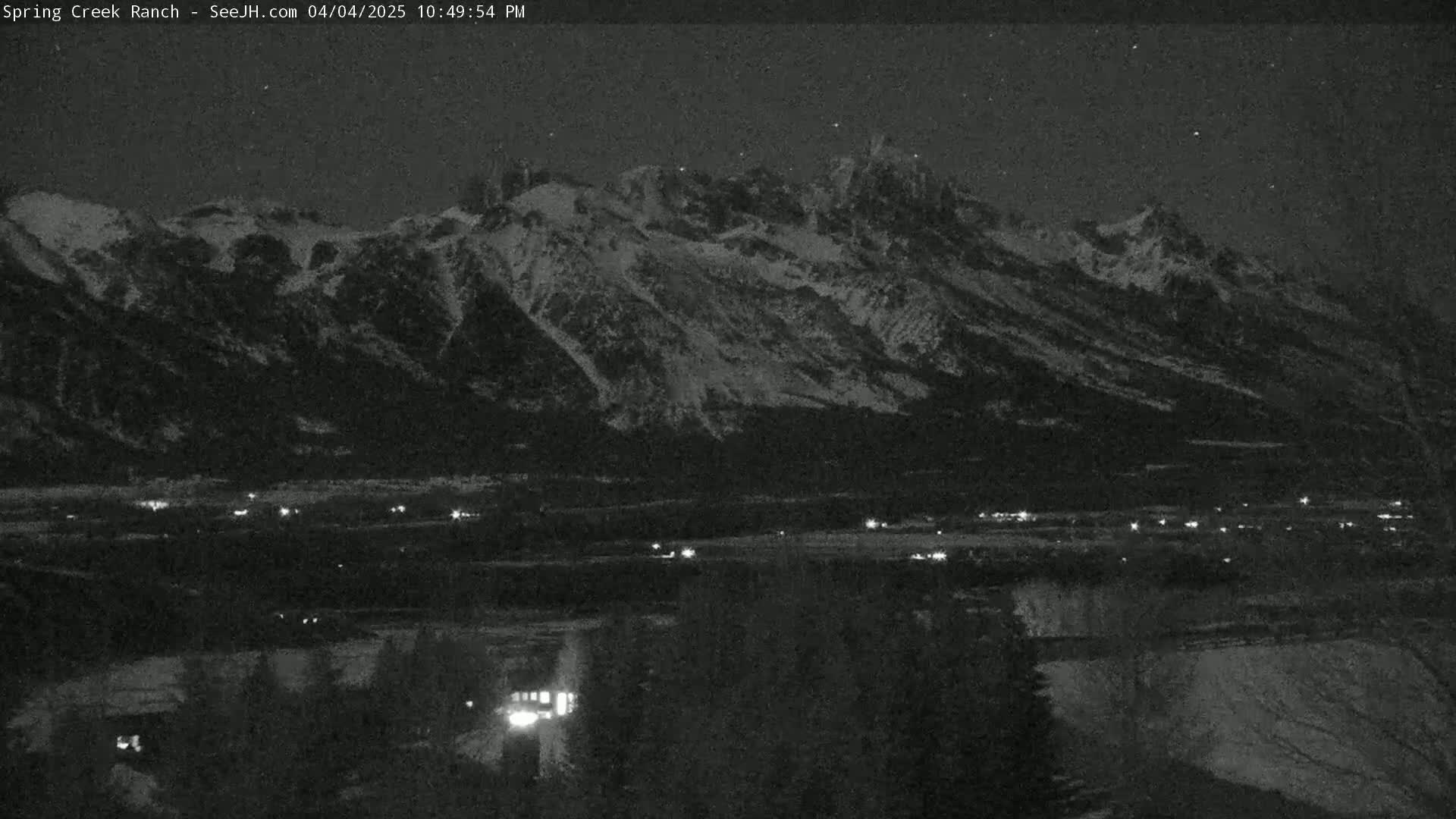 A nighttime, low-light view shows a snow-covered mountain range overlooking a valley with scattered lights below, under a clear, starry sky.