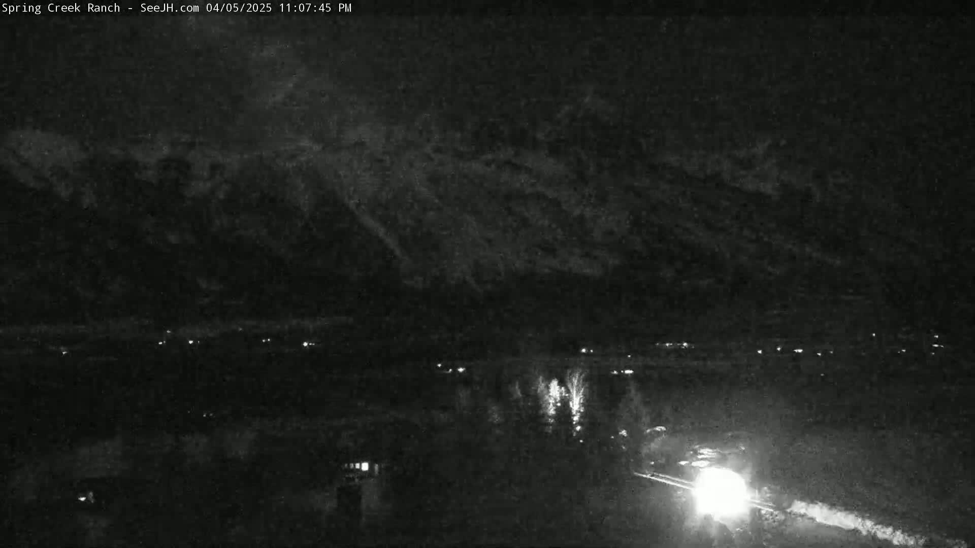 A nighttime, low-light view shows a dark mountain range under a cloudy sky, with a few scattered lights visible in a valley below and a bright light source, possibly headlights, moving along a road in the foreground.