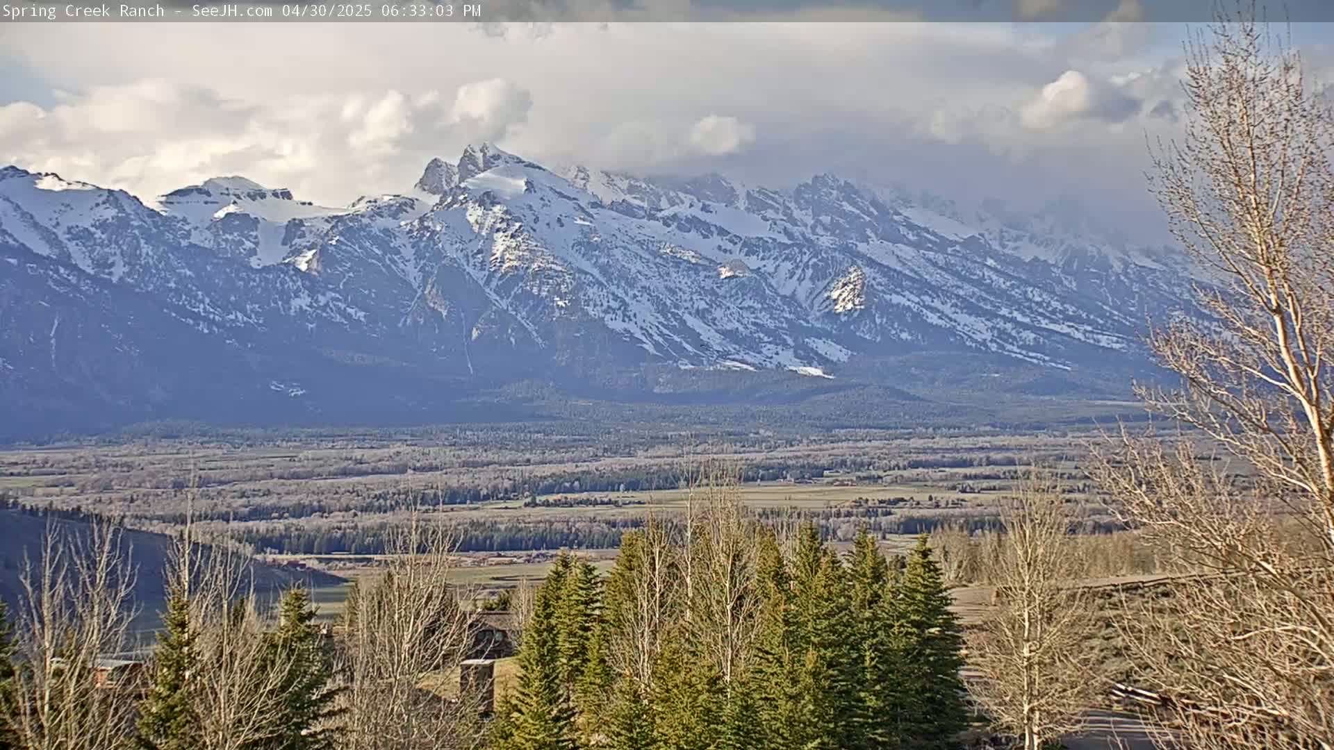 A partly cloudy day reveals a snow-capped mountain range in the distance, overlooking a valley of trees and shrubs.