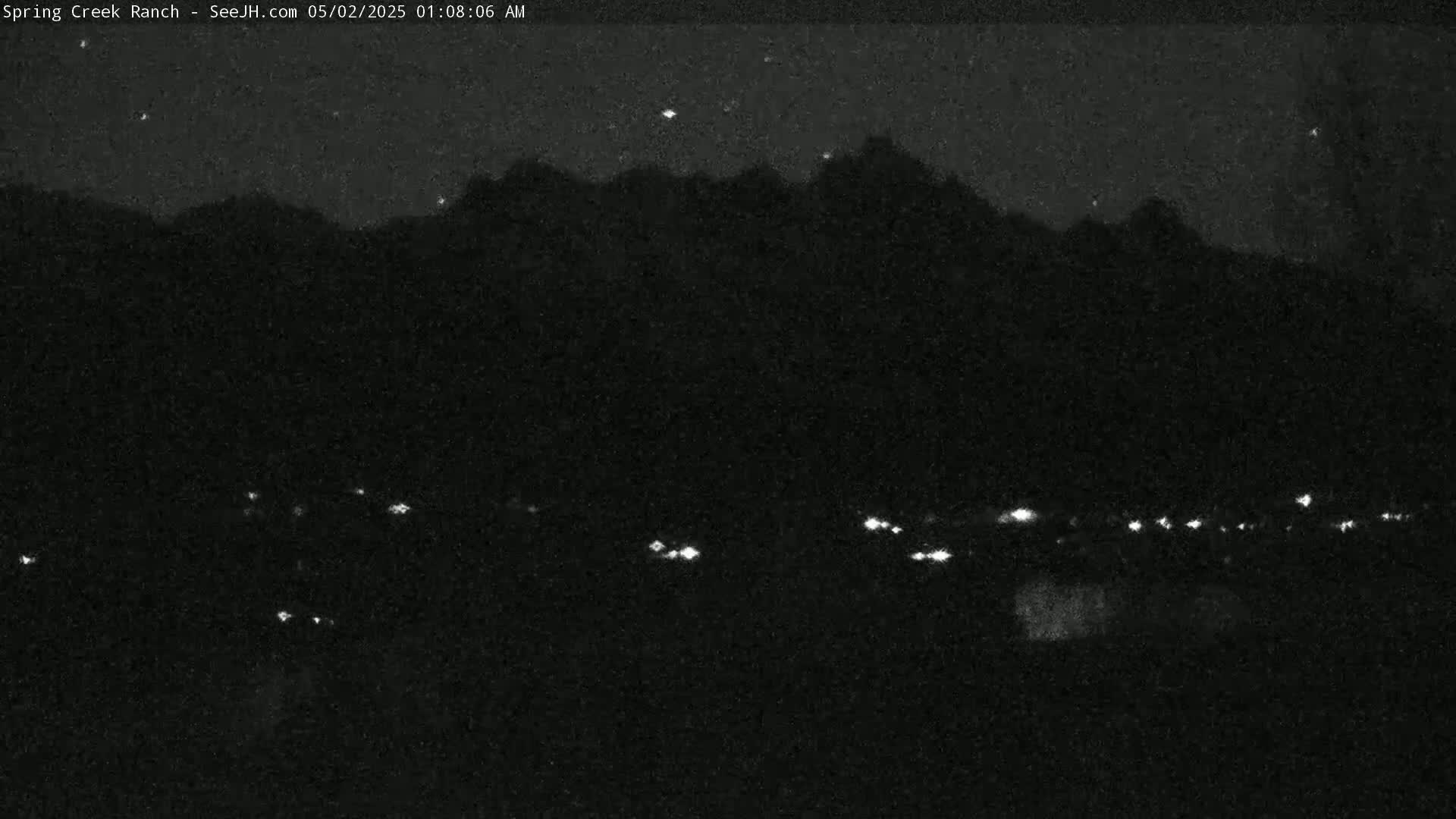 A nighttime, low-light view shows a dark mountain range silhouetted against a dark sky with a few stars visible, and a line of distant lights at the bottom.
