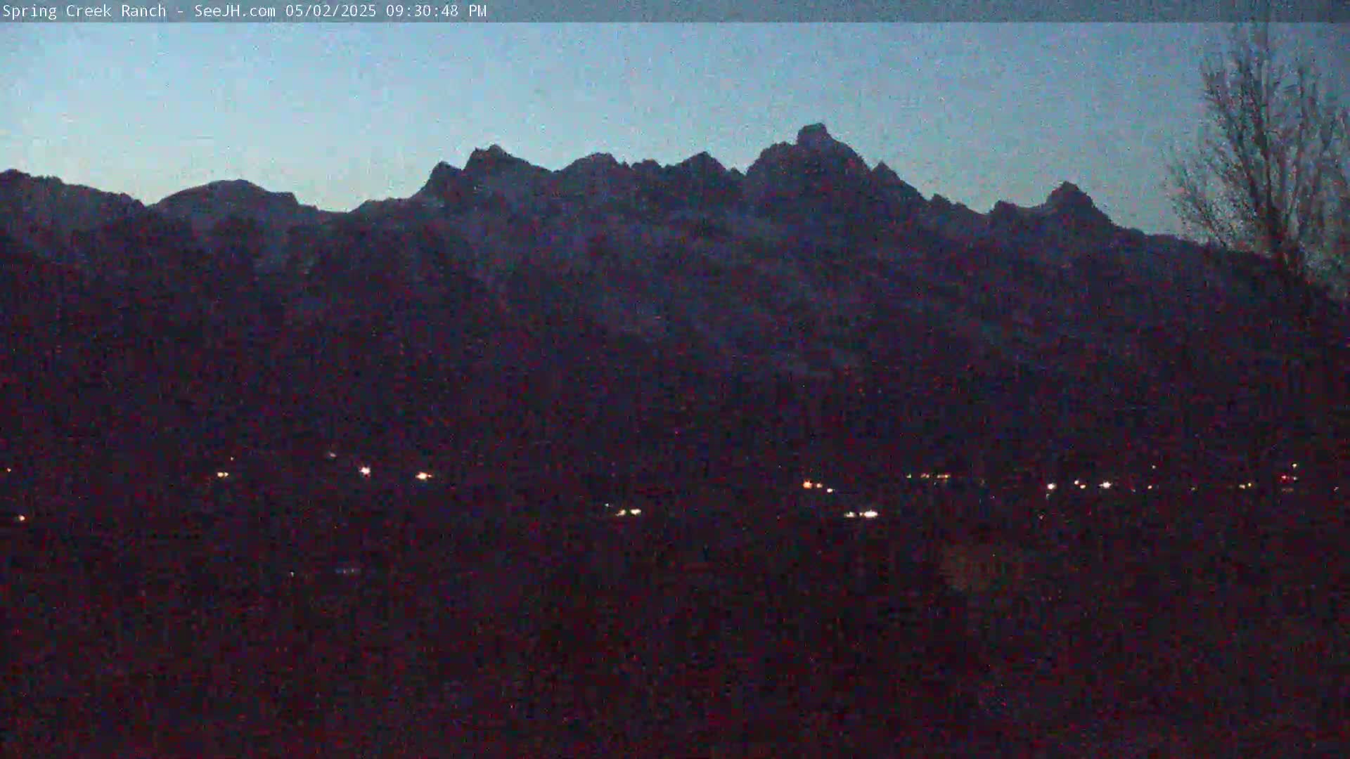 A dark, twilight view shows a mountain range silhouetted against a dusky blue sky, with a few faint lights visible in the foreground.