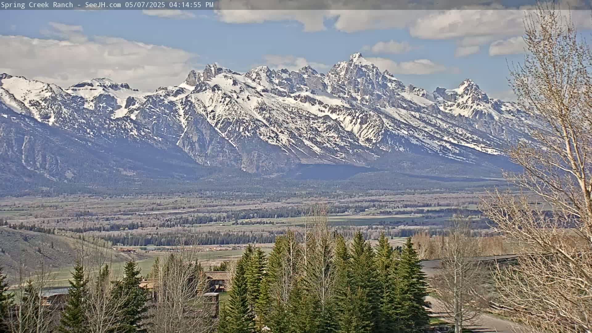 A partly cloudy day reveals a snow-capped mountain range in the distance, with a valley and evergreen trees in the foreground.