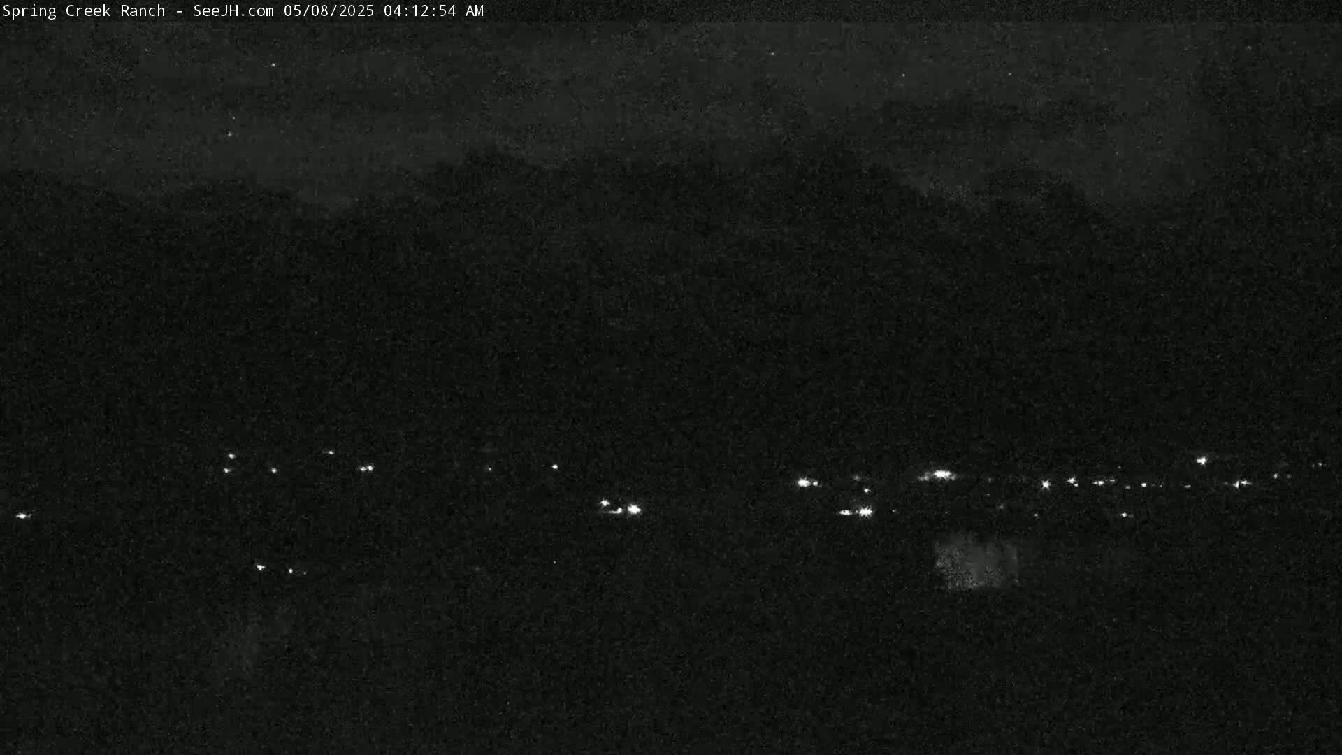 A nighttime, low-light view shows a distant line of small, faint lights against a dark, possibly cloudy sky and silhouetted mountains.
