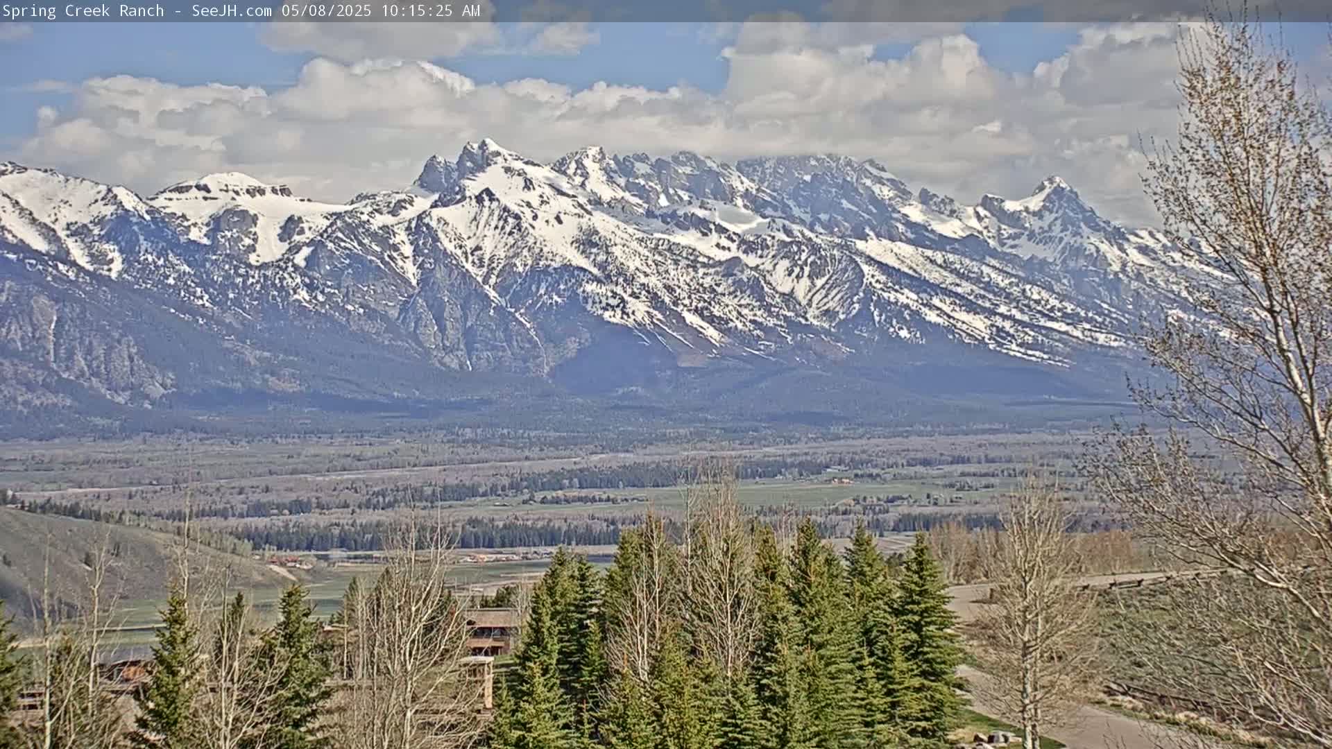A partly cloudy day reveals a snow-capped mountain range in the distance, overlooking a valley with trees and buildings in the foreground.