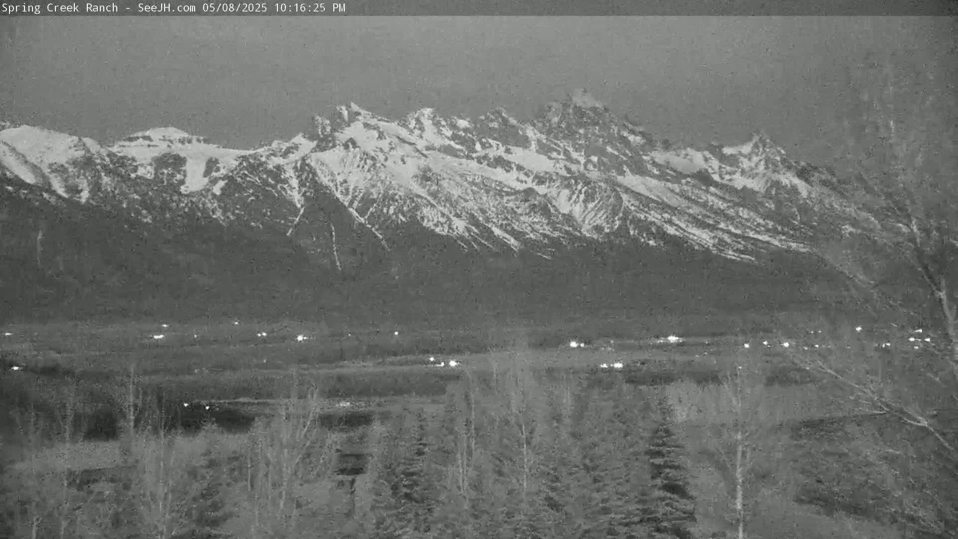 Snow-capped mountains are visible in the distance at twilight, with a flat area of sparse trees and faint lights in the foreground.