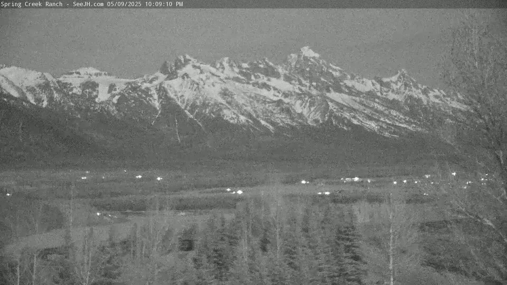 A snow-capped mountain range is visible in the distance, with a valley containing a few scattered lights below, under a dark, possibly twilight sky.