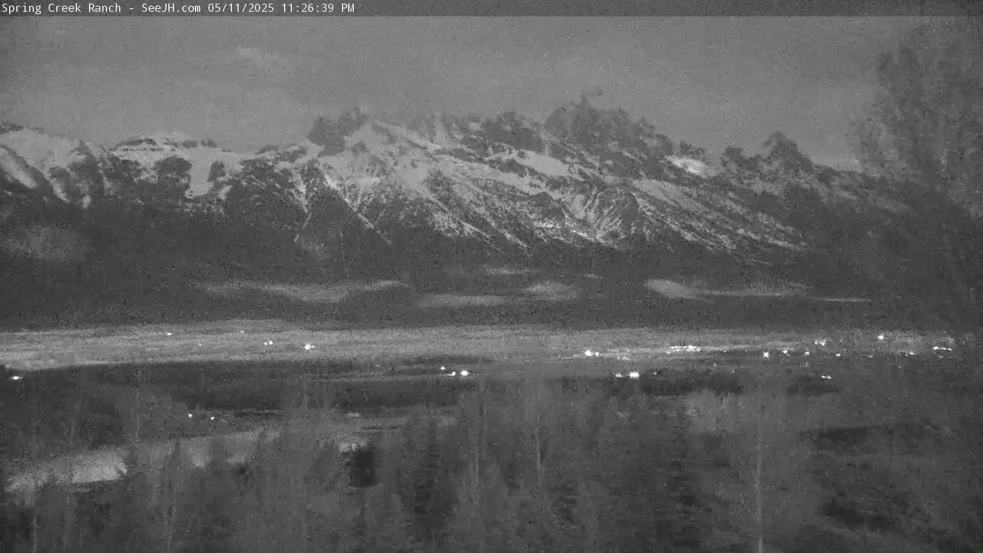 A nighttime, low-light view shows snow-capped mountains in the distance, with a few scattered lights visible in the valley below.