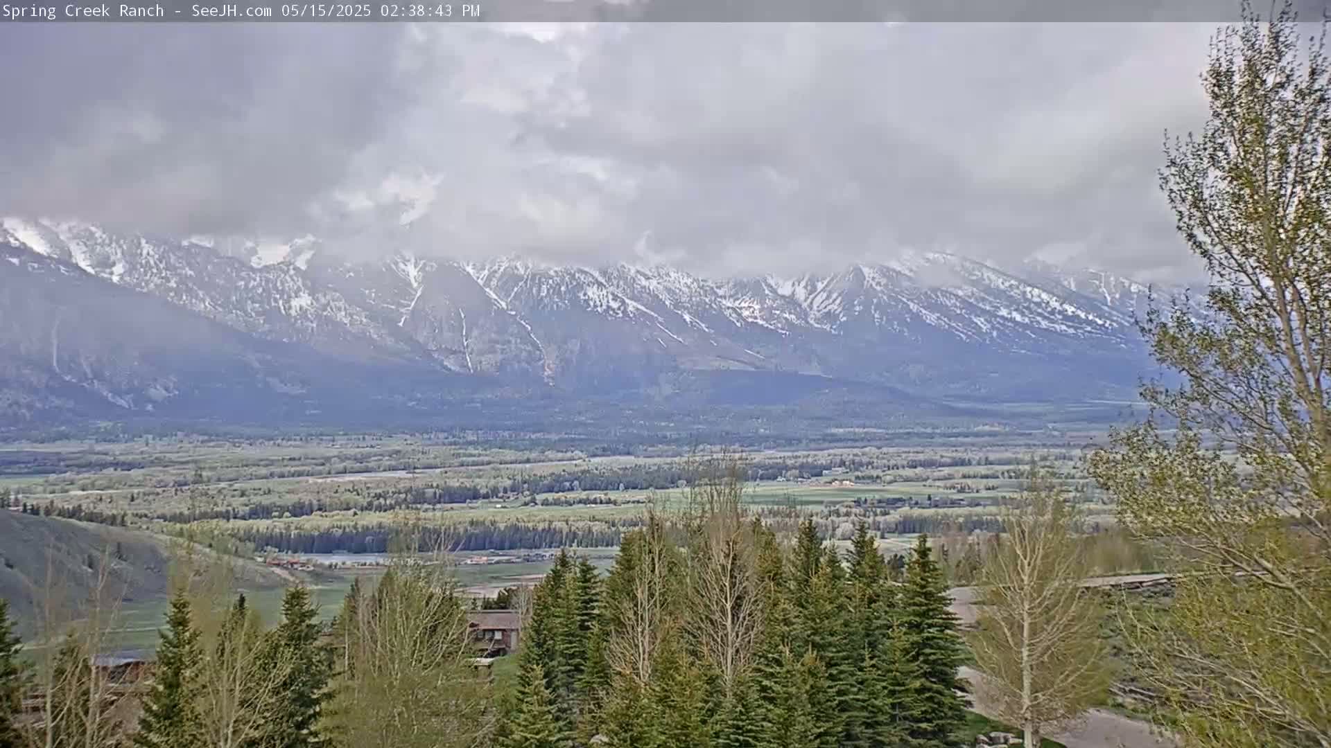 A long-range view shows snow-capped mountains under a cloudy sky, overlooking a valley of green fields and evergreens.