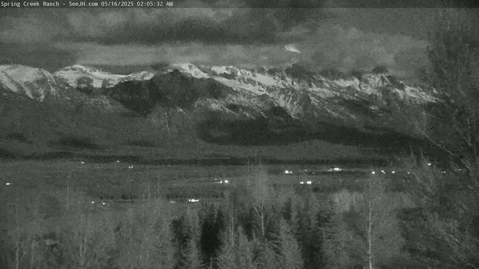 A nighttime view shows snow-capped mountains in the distance, with a flat valley and sparse trees in the foreground under a cloudy sky.