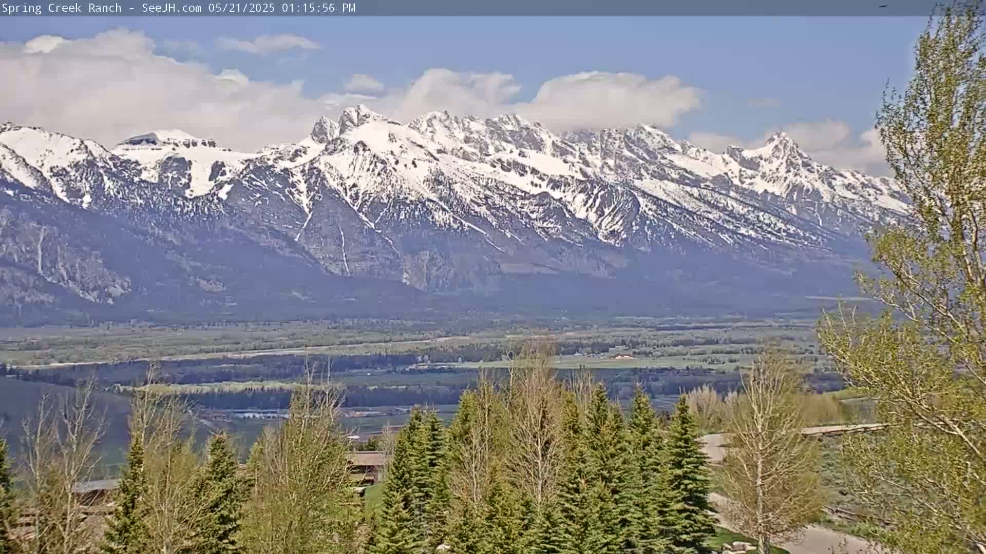 A partly cloudy day reveals a snow-capped mountain range in the distance, with a valley of green trees and fields in the foreground.
