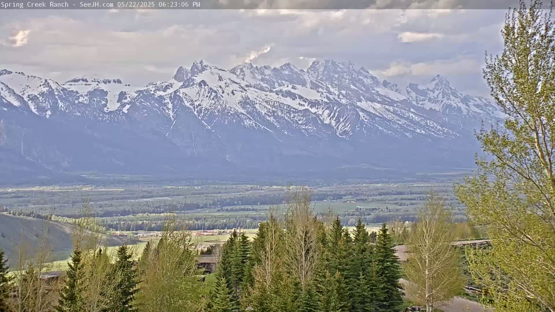 A snow-capped mountain range is visible in the distance, overlooking a valley with green trees and fields under a partly cloudy sky.