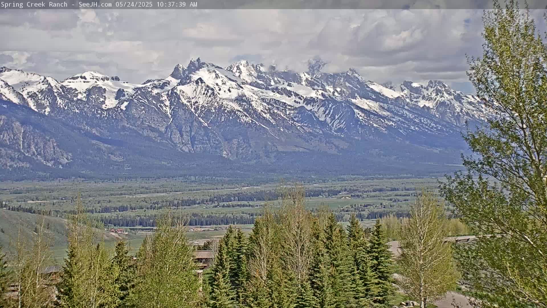 A partly cloudy day reveals a snow-capped mountain range in the distance, overlooking a valley with green fields and trees in the foreground.