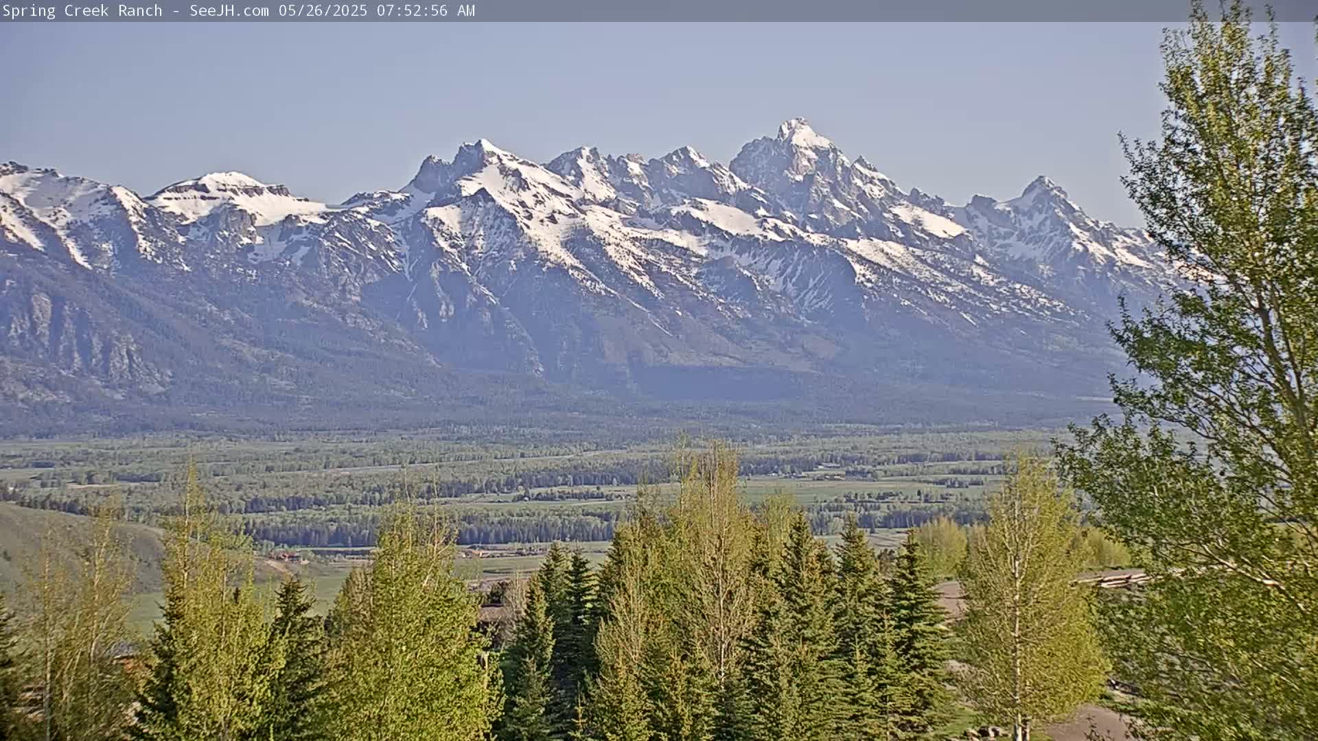 A snow-capped mountain range is visible in the distance under a clear sky, with a valley of green trees and fields in the foreground.