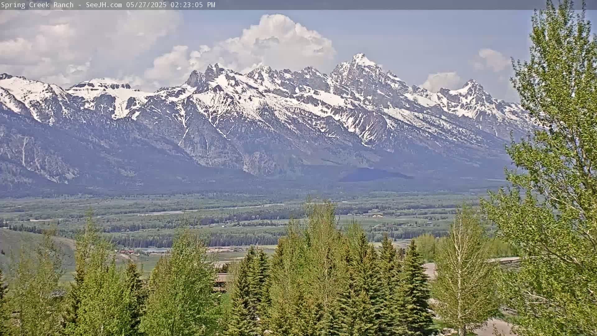 A partly cloudy day reveals a snow-capped mountain range in the distance, overlooking a valley filled with green vegetation and trees in the foreground.