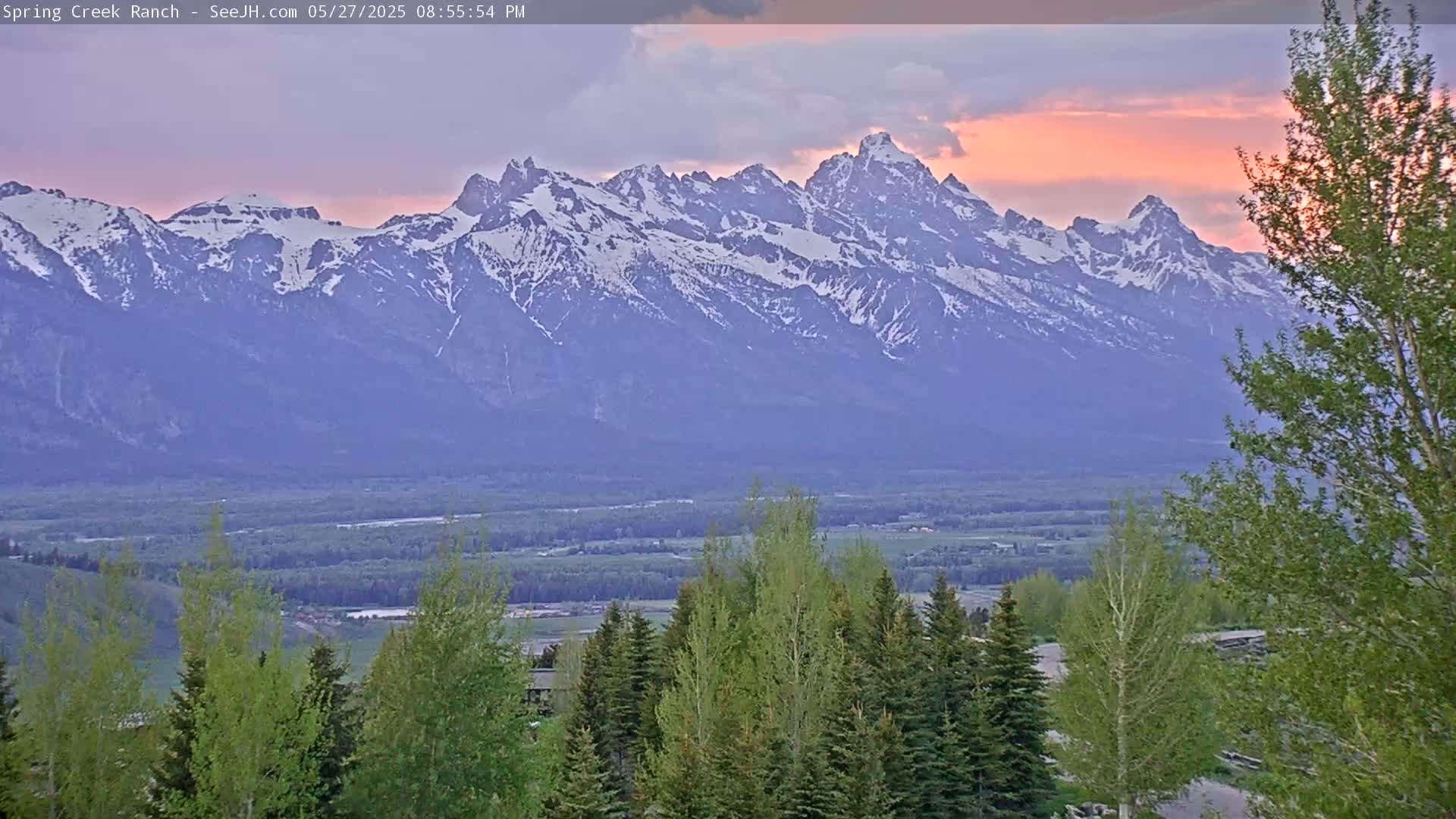 A snow-capped mountain range is visible at sunset, with a valley of green trees and land in the foreground.