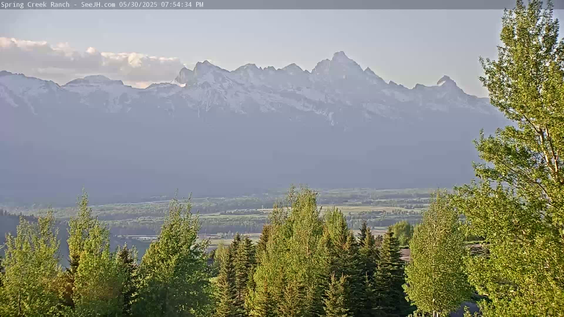 A hazy, partly cloudy day reveals a snow-capped mountain range in the distance, with a valley and green trees in the foreground.