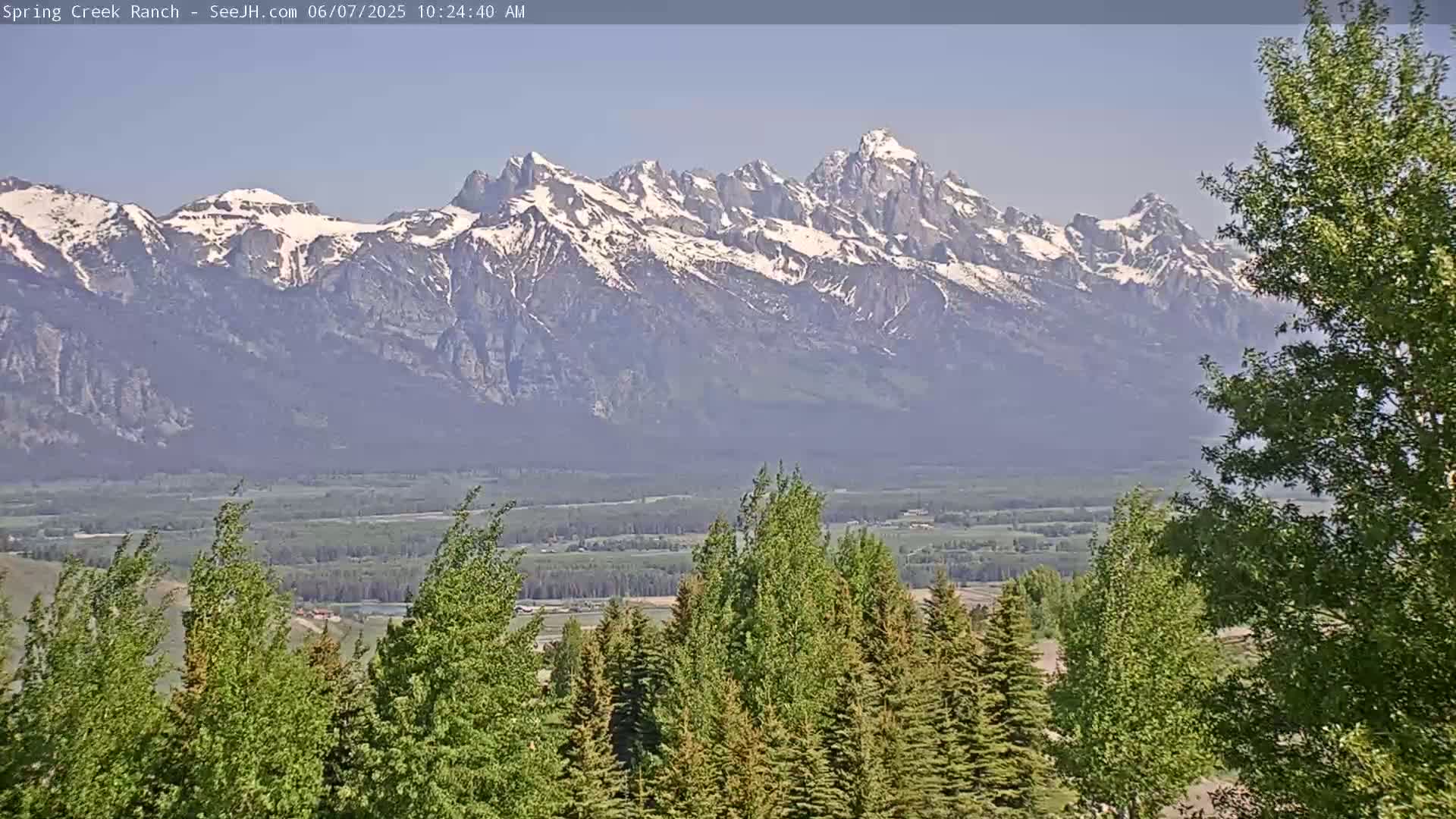 A snow-capped mountain range is visible in the distance, beyond a valley of green trees and fields under a clear sky.