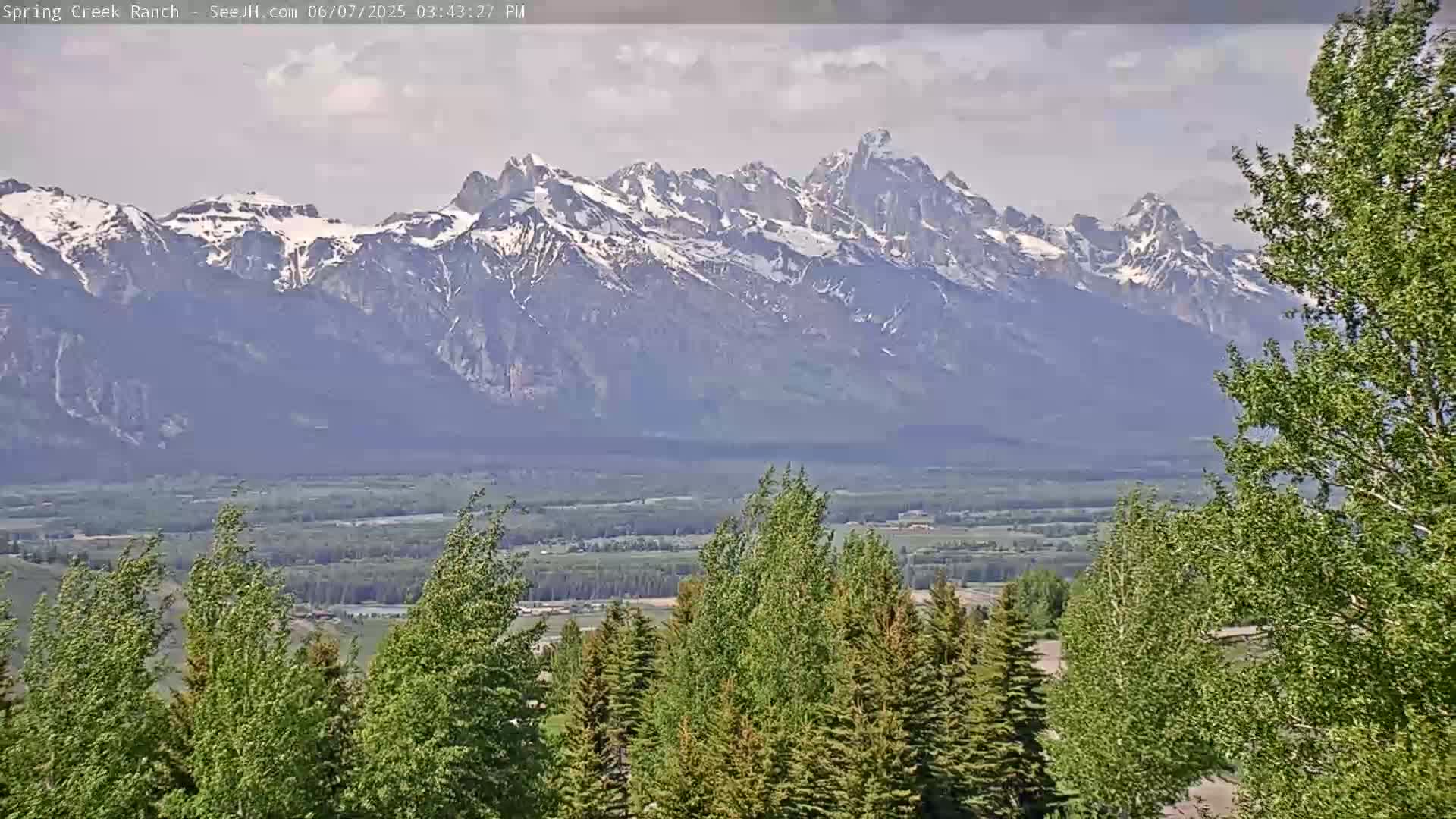 A partly cloudy day reveals snow-capped mountains in the distance, overlooking a valley of green trees and fields.