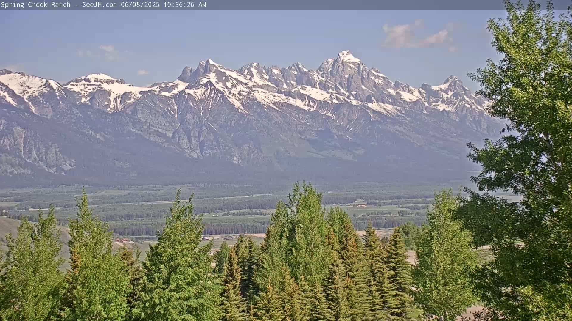 A snow-capped mountain range is visible in the distance, with a valley of green trees and fields in the foreground under a mostly clear sky.