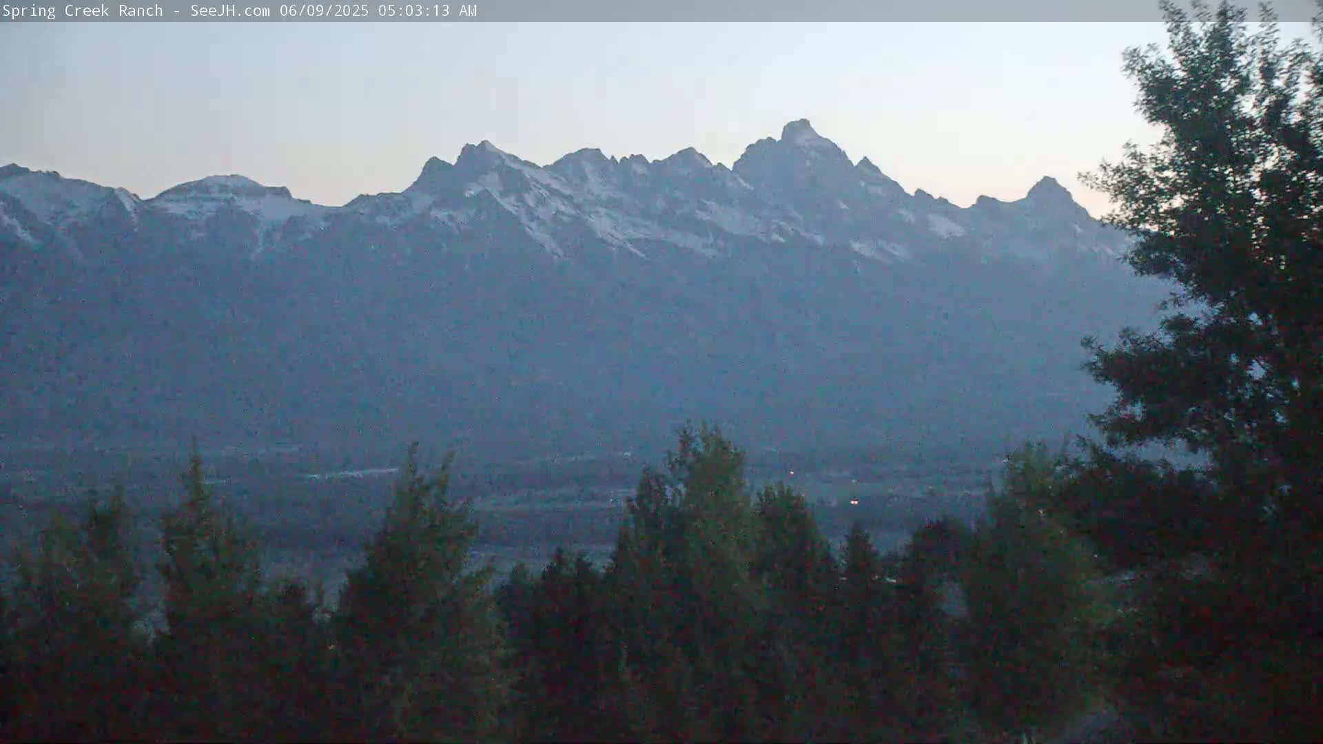 Snow-capped mountains are visible in the distance, with a valley of dark green trees in the foreground under a hazy, light sky at dawn.