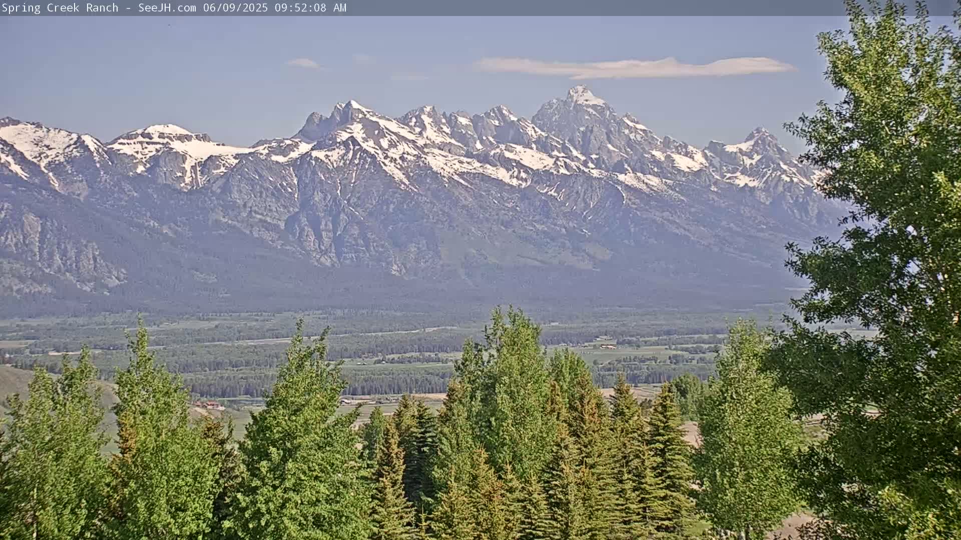 Snow-capped mountains are visible in the distance, overlooking a valley filled with green trees and under a mostly clear sky.