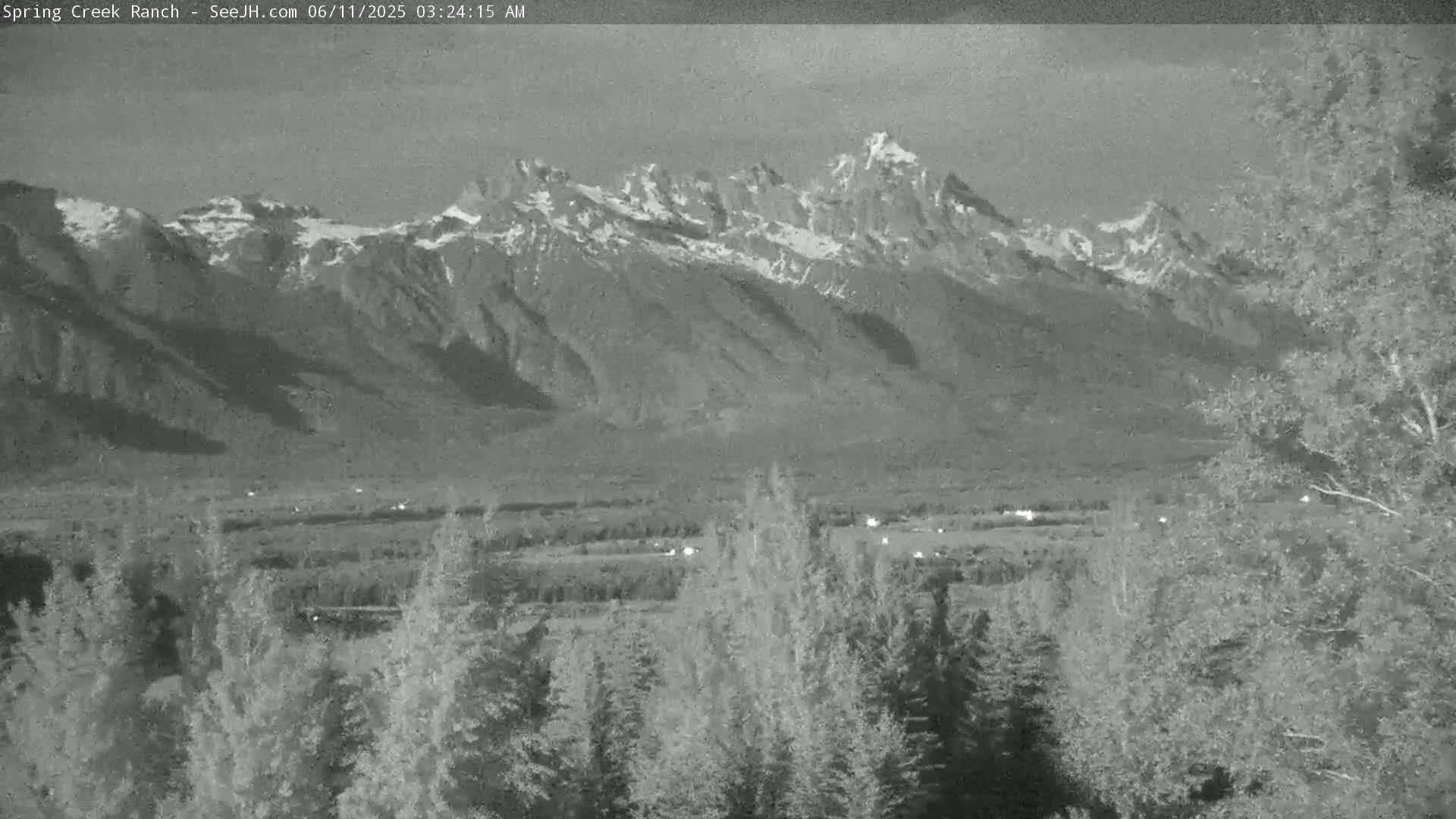 A nighttime, grayscale view shows snow-capped mountains in the distance, with a foreground of dark trees and a valley illuminated by faint lights.