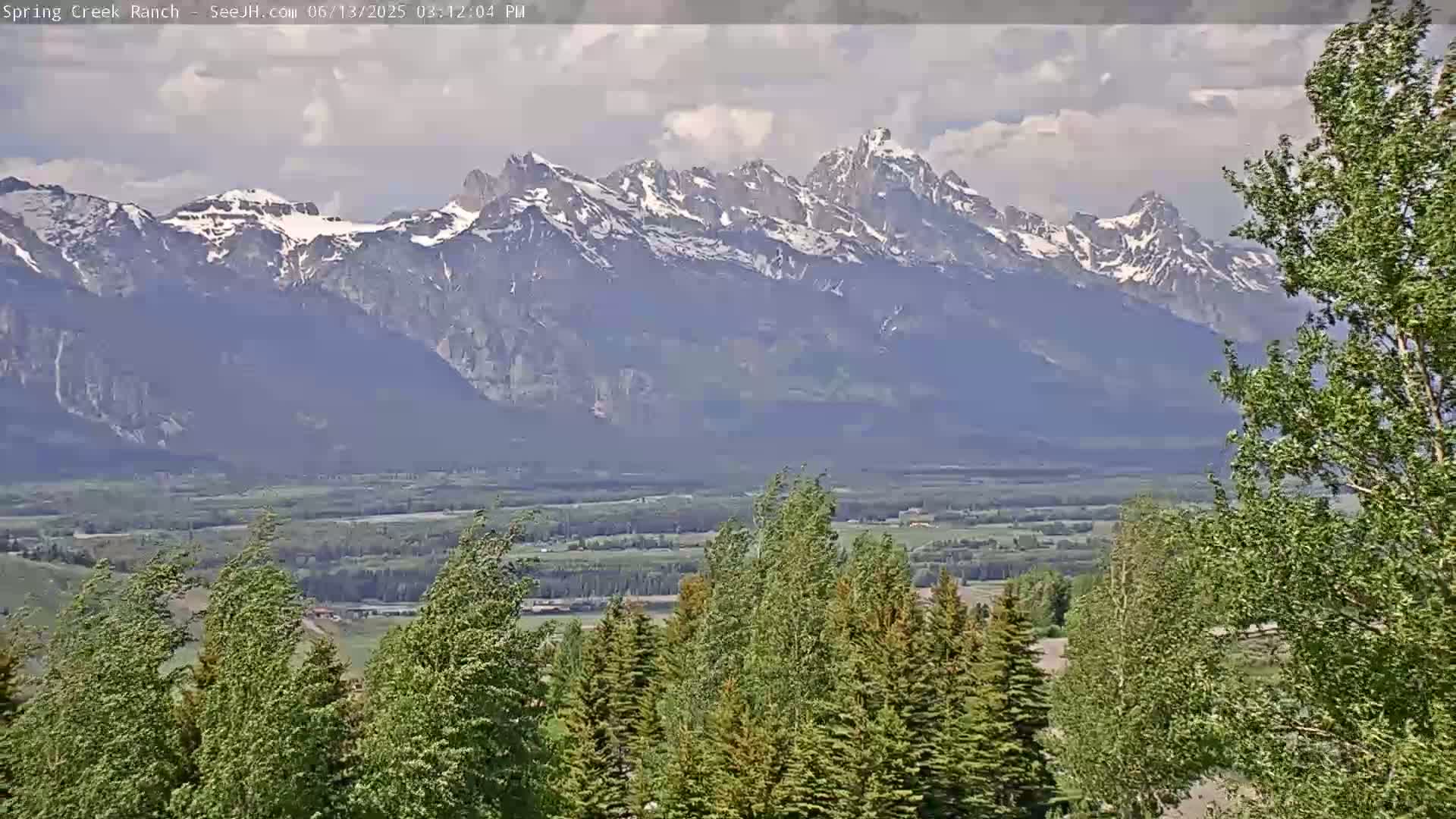 A partly cloudy day reveals a view of snow-capped mountains in the distance, with a valley of green trees and fields in the foreground, where the trees are blowing in the wind.