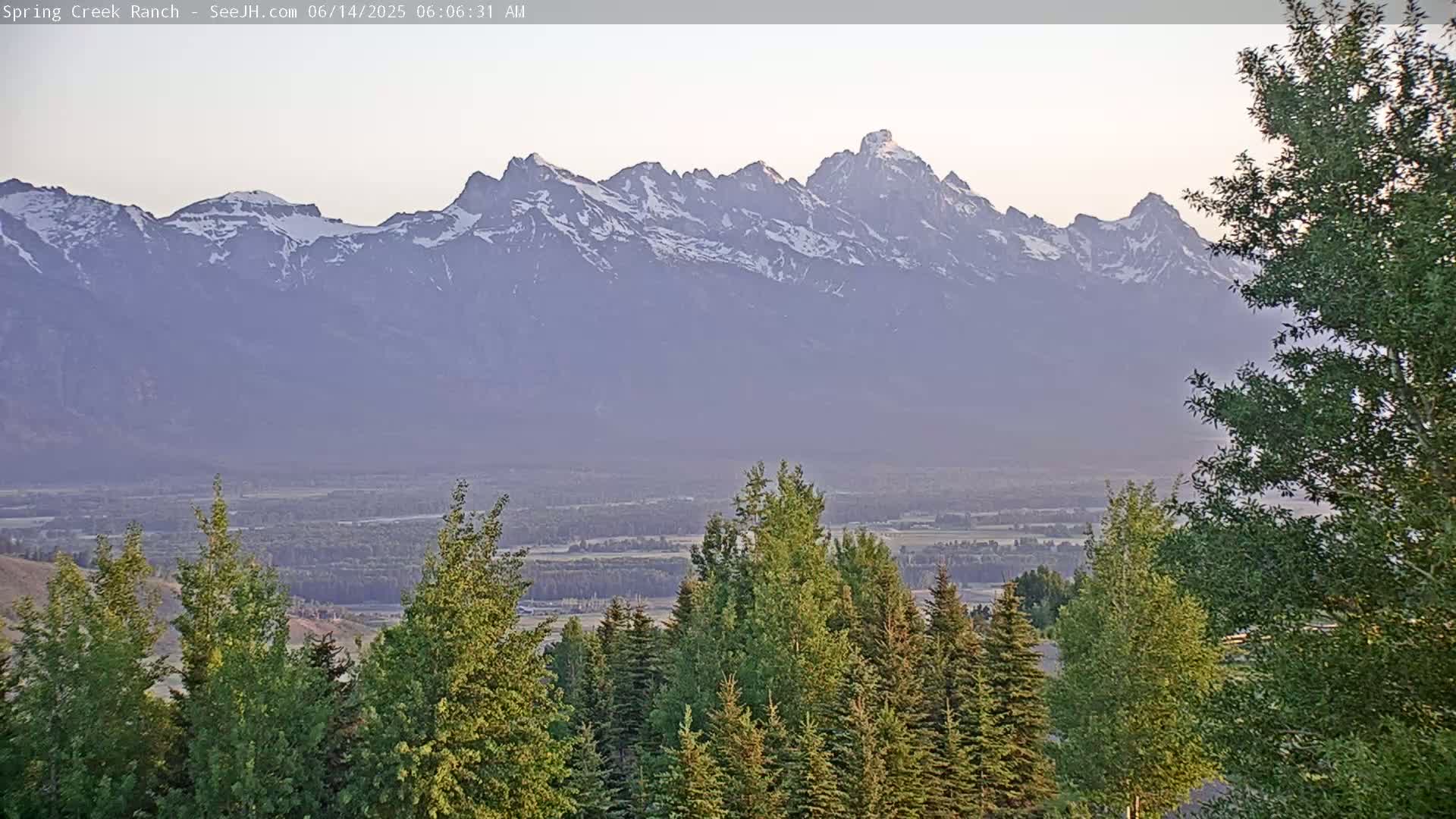A hazy, sunrise view shows a snow-capped mountain range in the distance, with a valley and evergreen trees in the foreground.