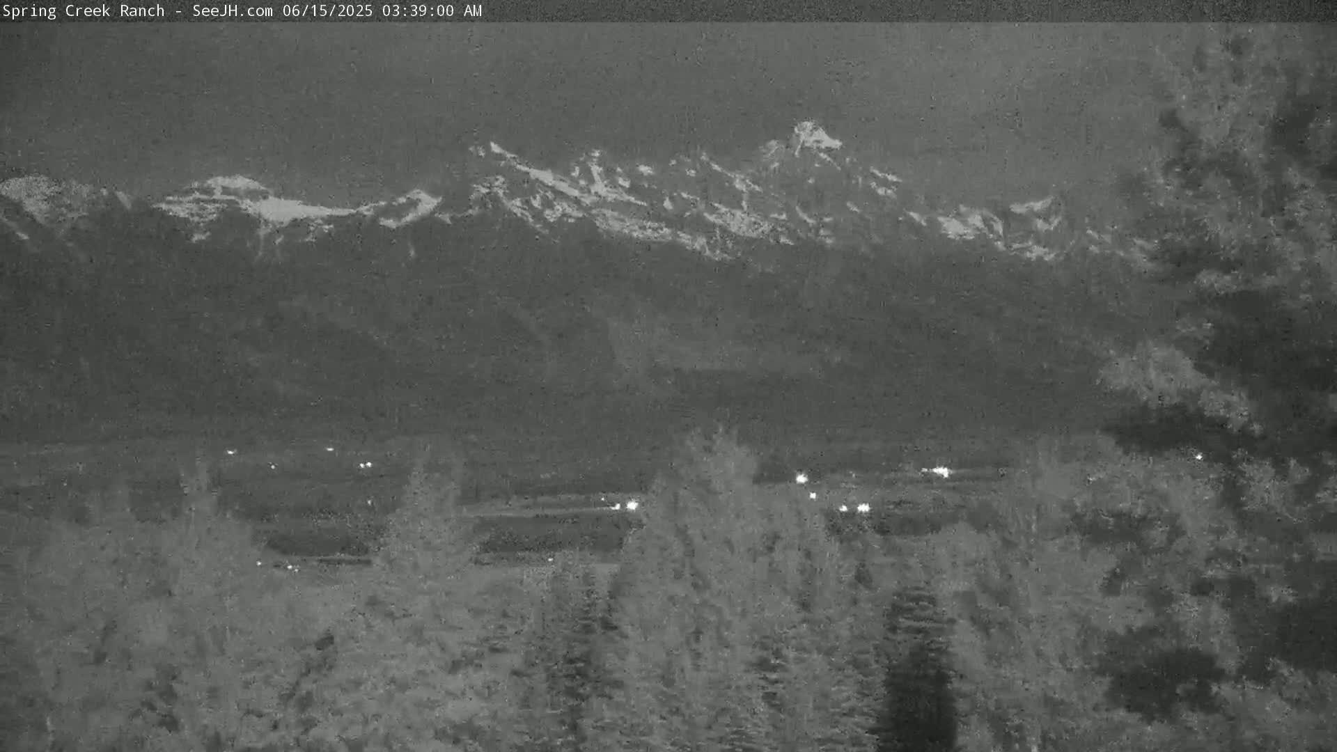 A nighttime view shows snow-capped mountains in the distance, with a valley and dark trees in the foreground under a dark, possibly overcast sky.