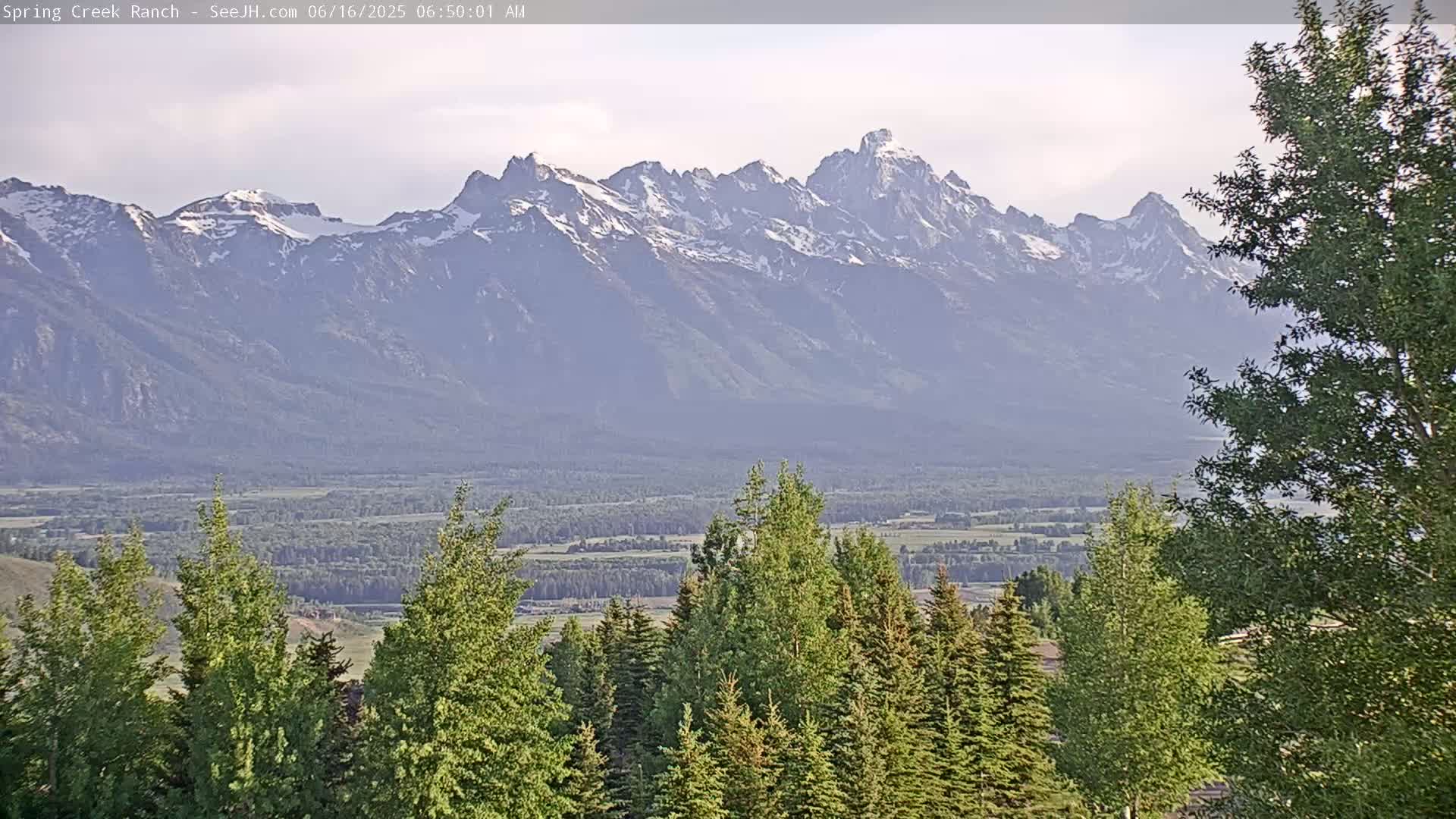 A mountain range capped with snow is visible in the distance, beyond a valley of green trees and fields under a hazy sky.