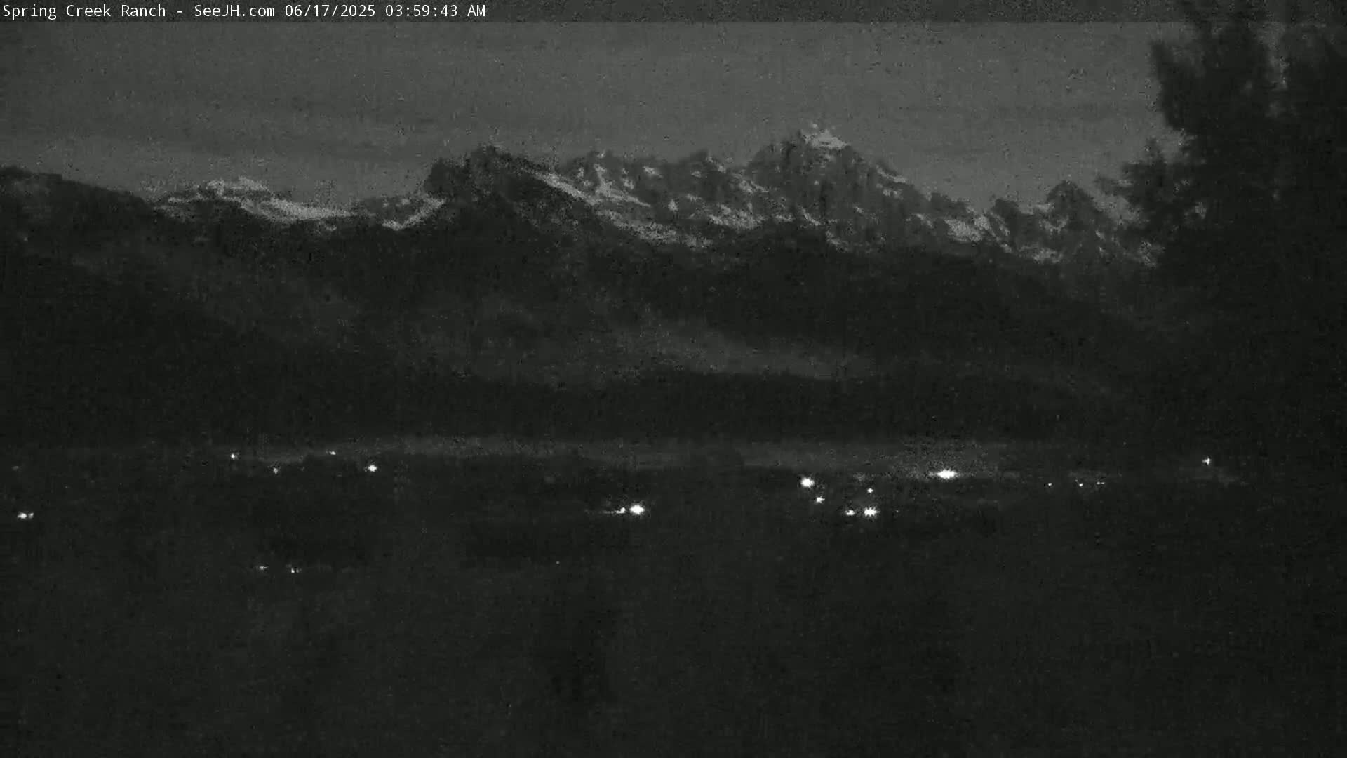 A nighttime, low-light view shows a snow-capped mountain range in the distance, with a few scattered lights visible in the foreground.