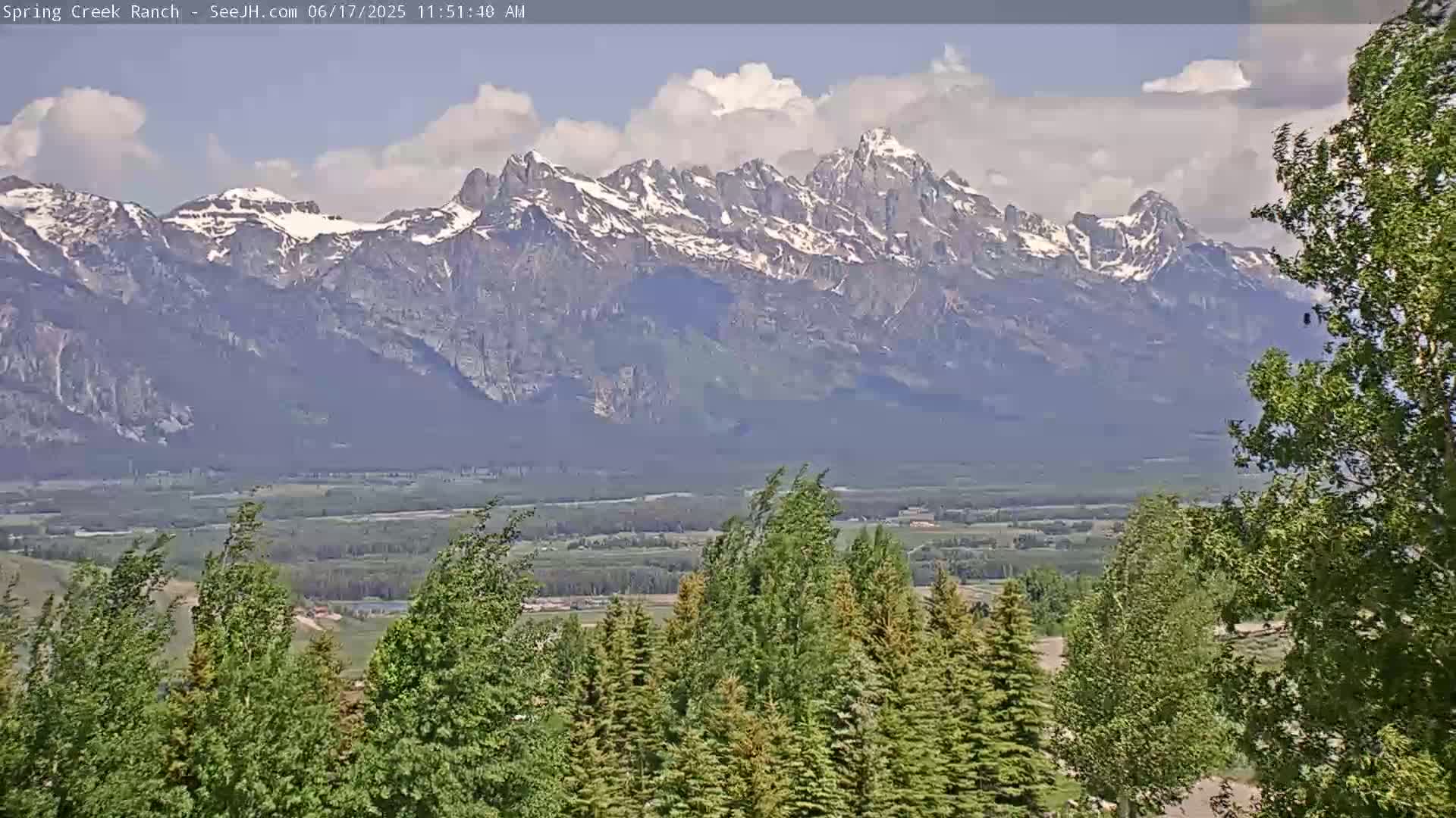 A partly cloudy day reveals a valley of green trees and fields, with a snow-capped mountain range in the background.
