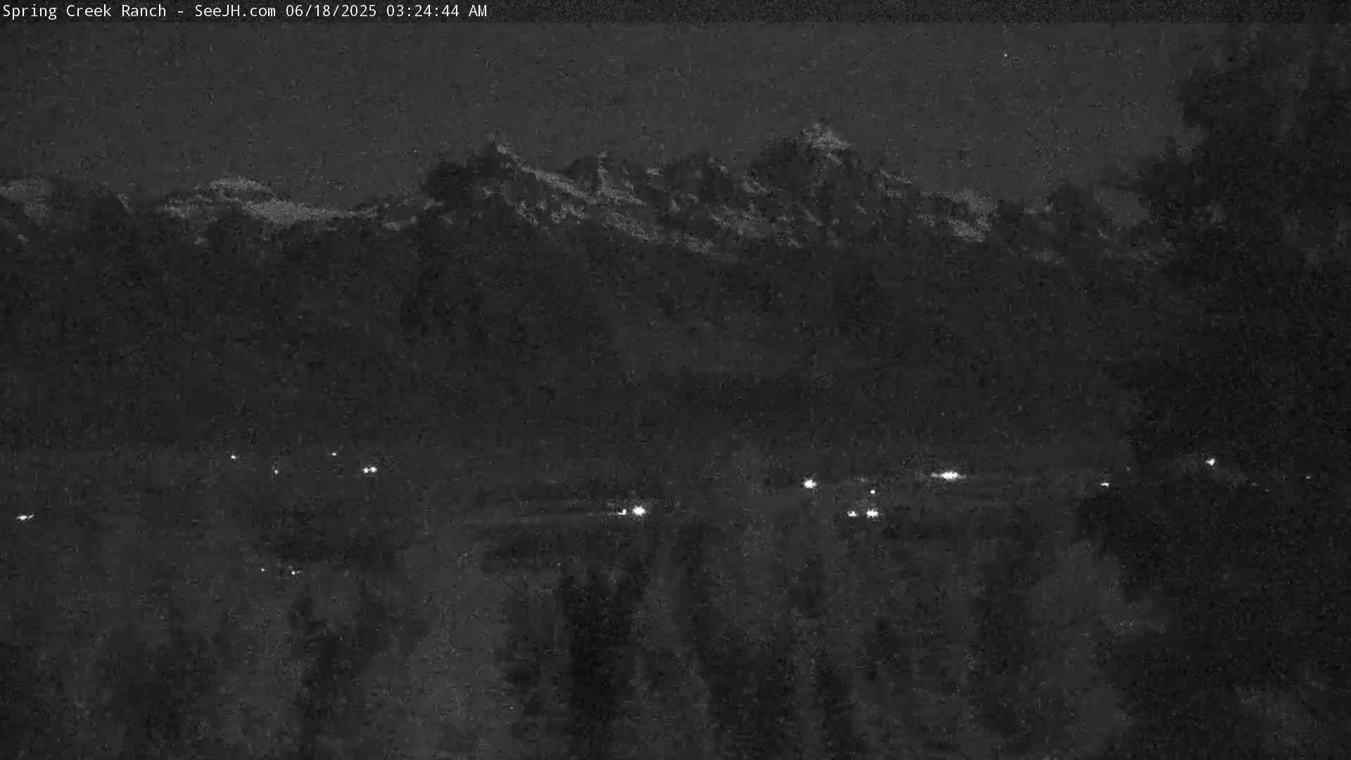 A nighttime long shot shows a mountain range in the distance, with a few scattered lights visible below in the foreground under a dark sky.