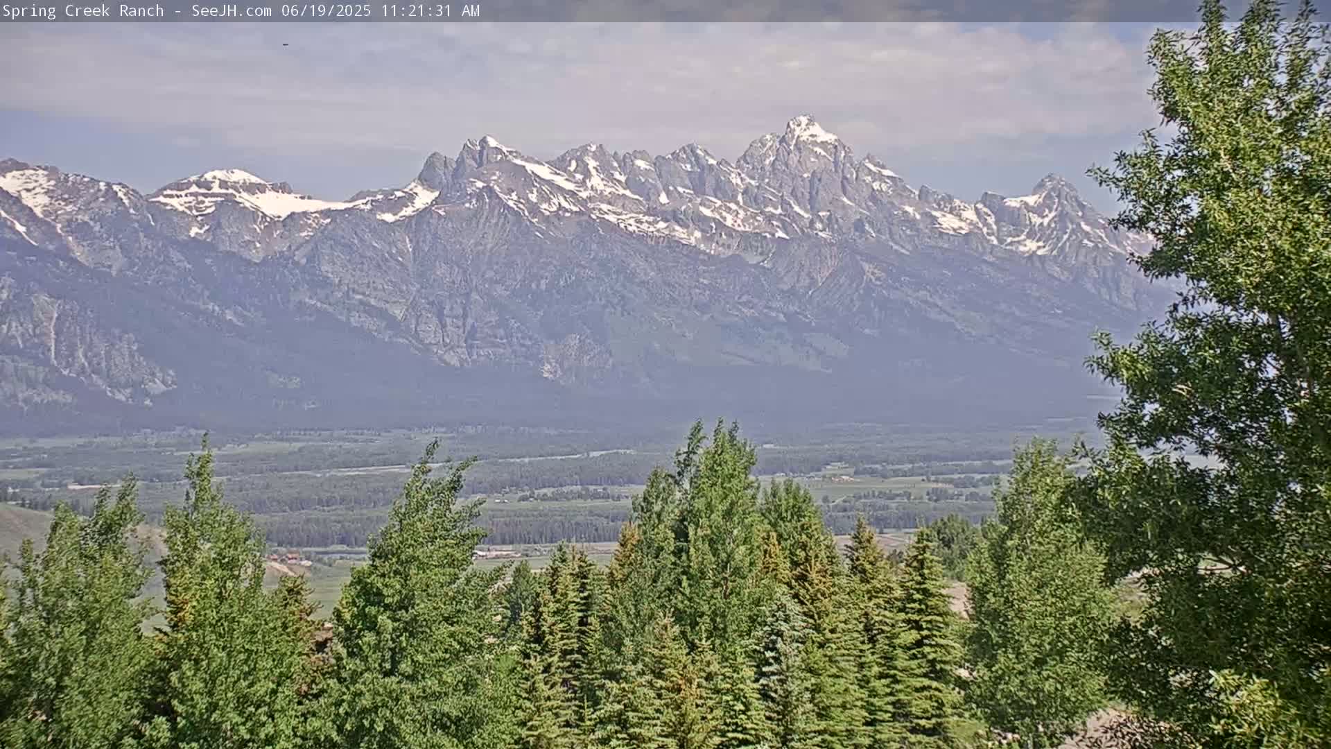 A snow-capped mountain range is visible in the distance, with a hazy valley and green trees in the foreground under partly cloudy skies.