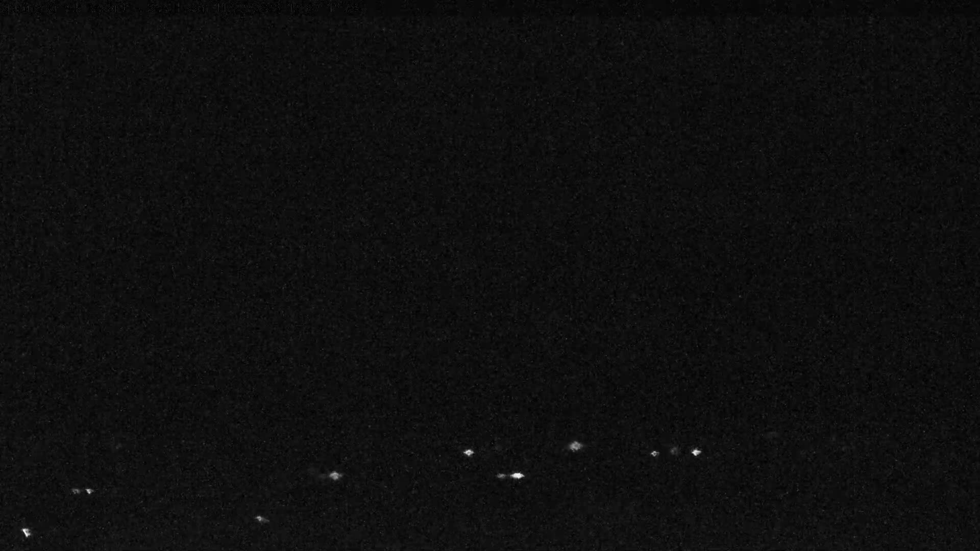 The image captures a very dark and grainy outdoor scene at night, revealing scattered small, bright white lights twinkling across the bottom portion, indicative of a clear night.