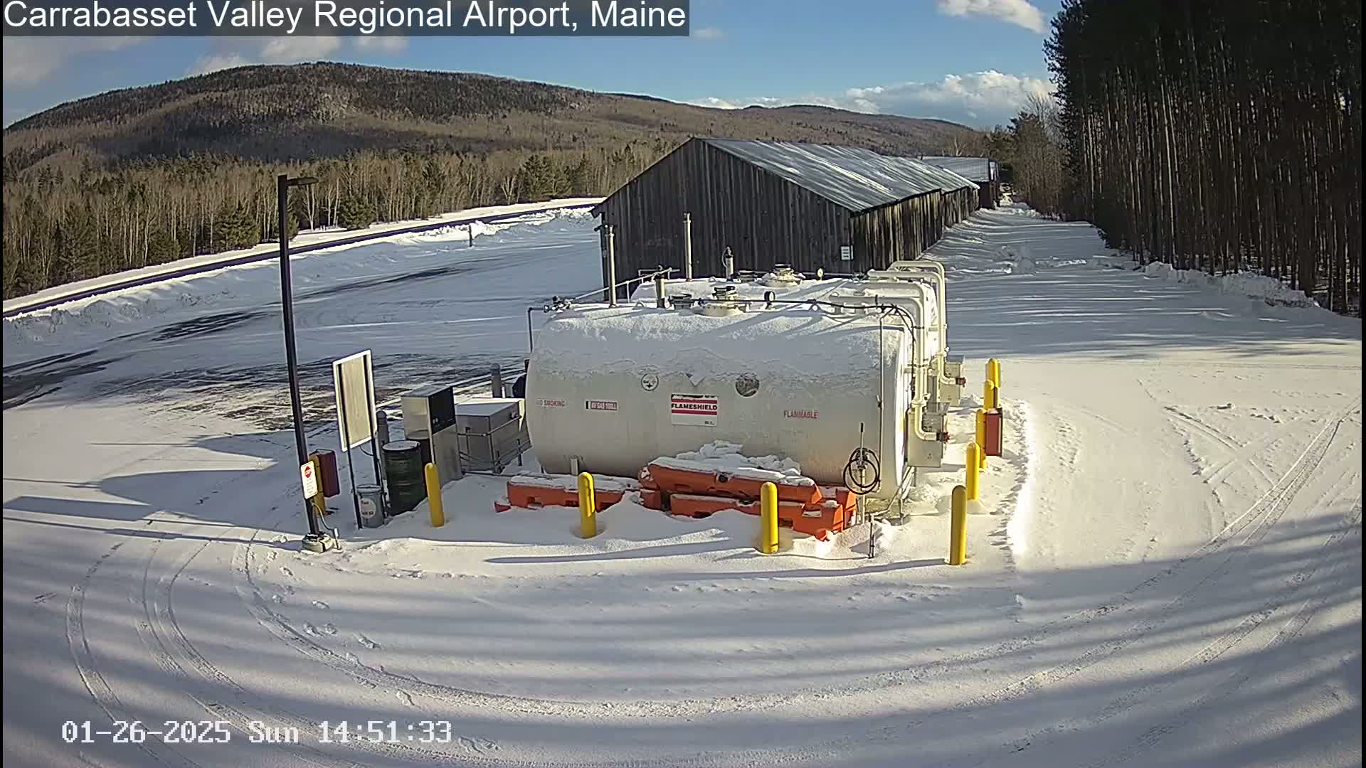 A snow-covered airport fuel depot is situated in a sunny, snowy landscape with a wooded hillside in the background.