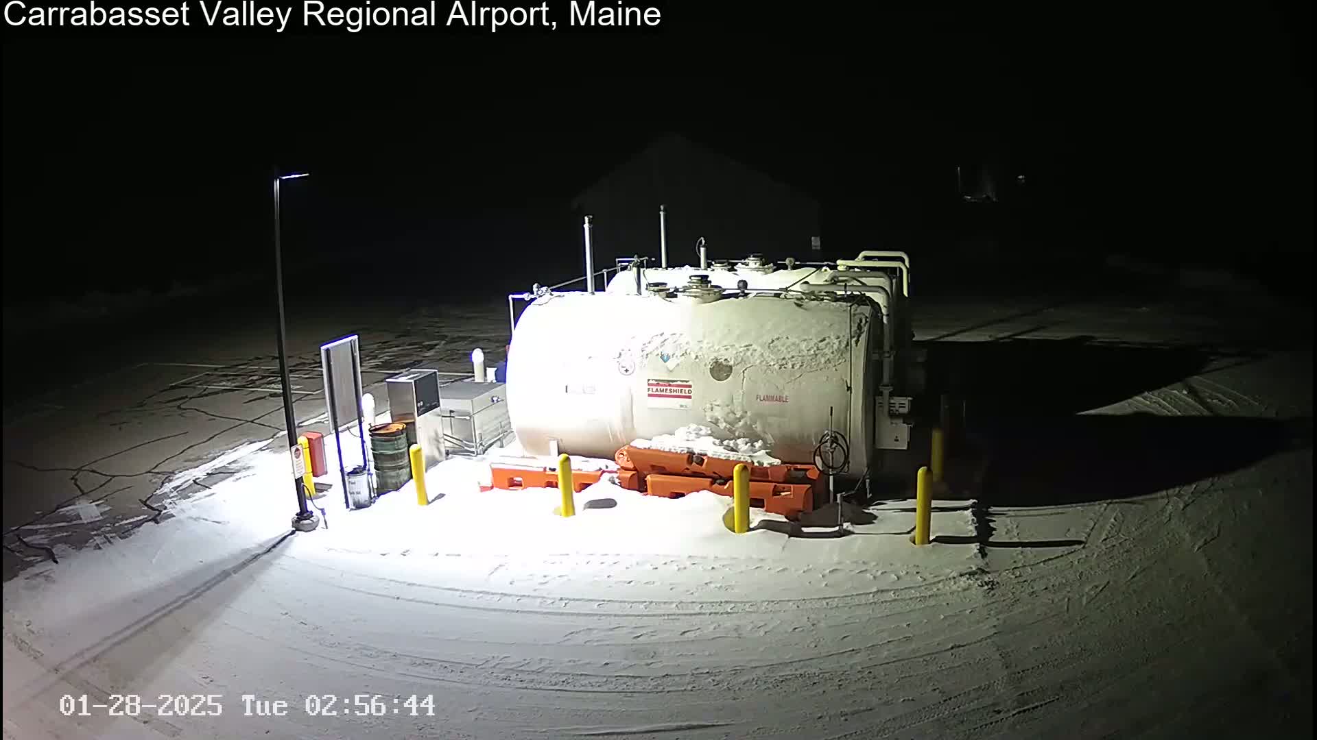 A snow-covered fuel tank and surrounding equipment are illuminated at night in a parking lot.