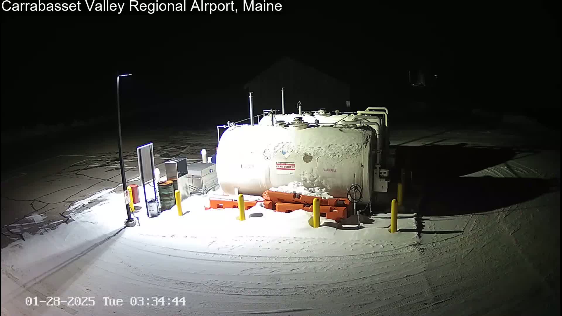 A large, snow-covered fuel tank sits in a snow-covered area at night, illuminated by a single light pole.