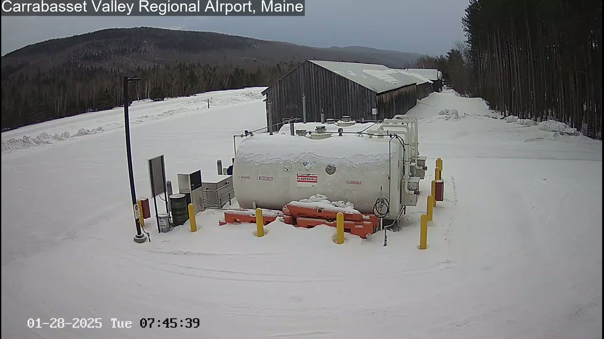 A large, snow-covered fuel tank sits in a snowy, outdoor area near a wooden building and a stand of trees under an overcast sky.