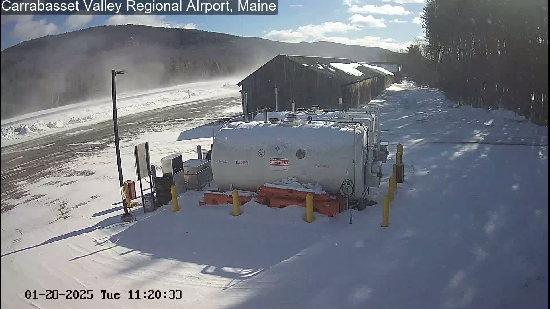 A large, snow-covered fuel tank sits in a snowy airport lot with a wood building and a snow-covered road in the background, under a partly sunny sky.