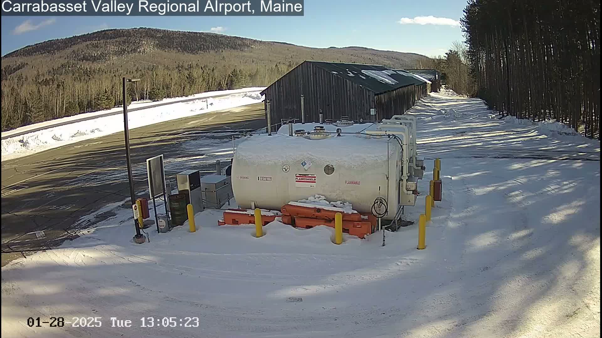 A large, white, flammable storage tank sits in a snow-covered area next to a long, dark building, with trees and a mountain visible in the background under a sunny sky.