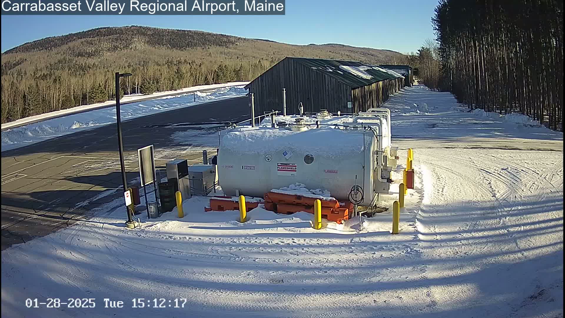 A snow-covered airport fuel station sits in a parking lot next to a long building under a clear, sunny sky.