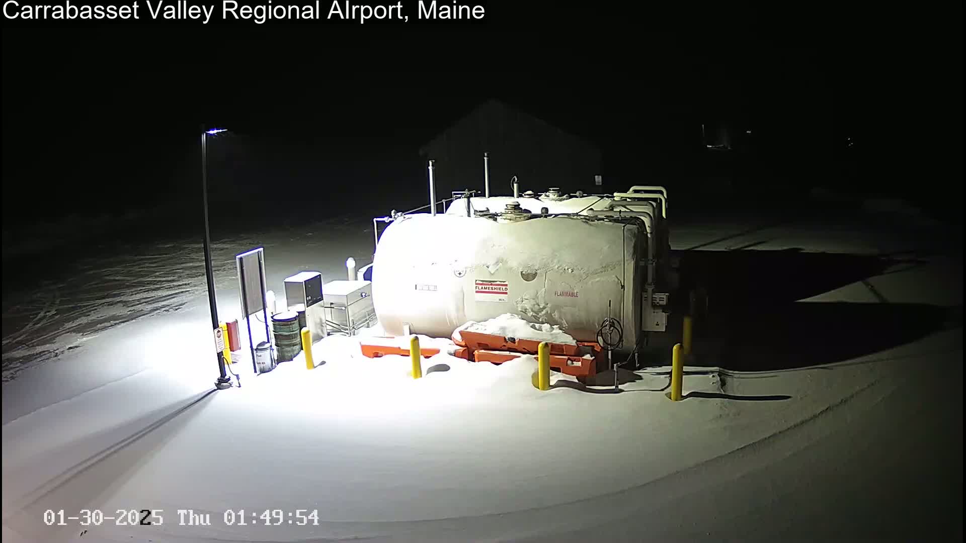 A large, snow-covered fuel tank sits in a snow-covered area at night, illuminated by a nearby lamppost.