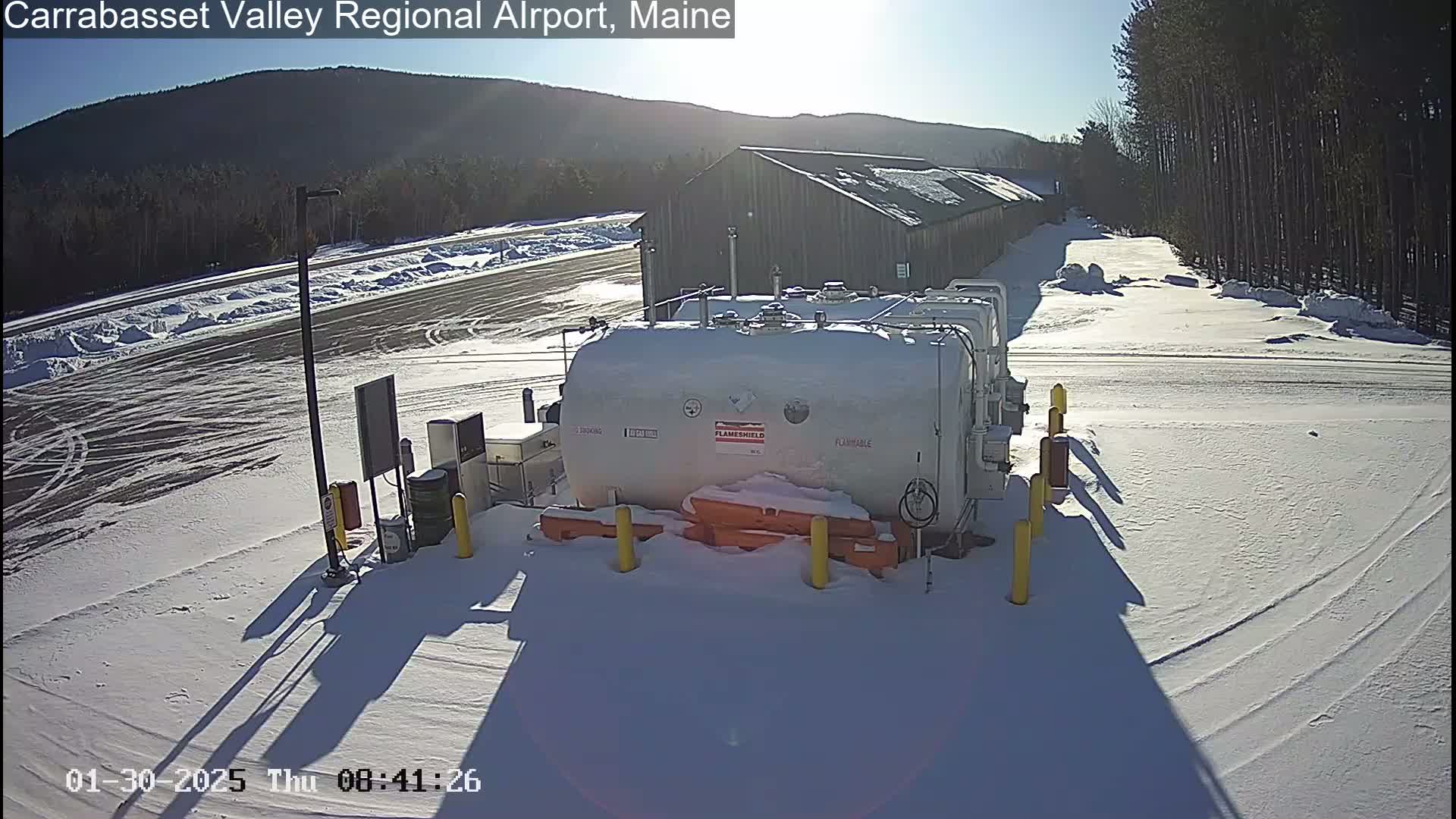 A snowy airfield with a large fuel tank and a long, low building is visible under a sunny sky.