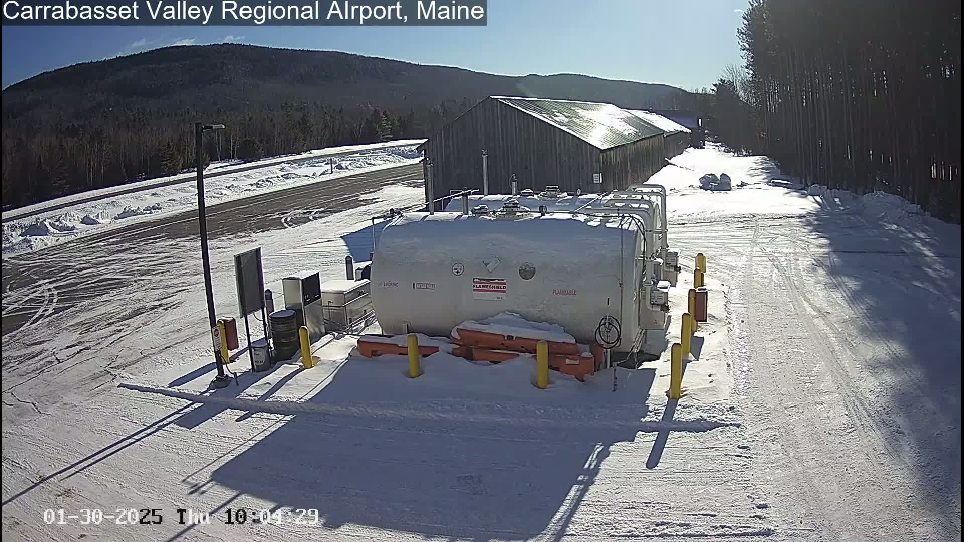 A snowy airfield with several large, white fuel tanks sits near a long, wooden building under a bright, sunny sky.