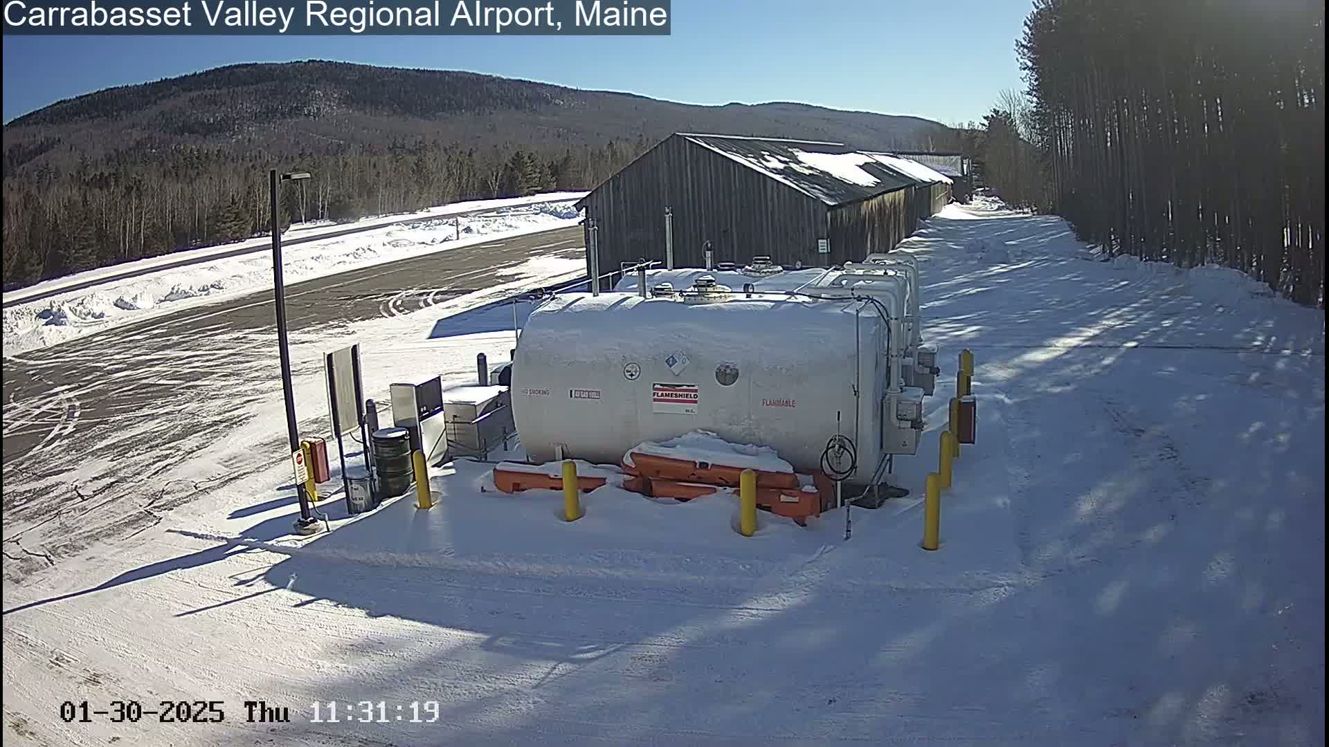 A large white fuel tank sits in a snow-covered airport lot on a sunny day, near a long wooden building and a snow-covered roadway.